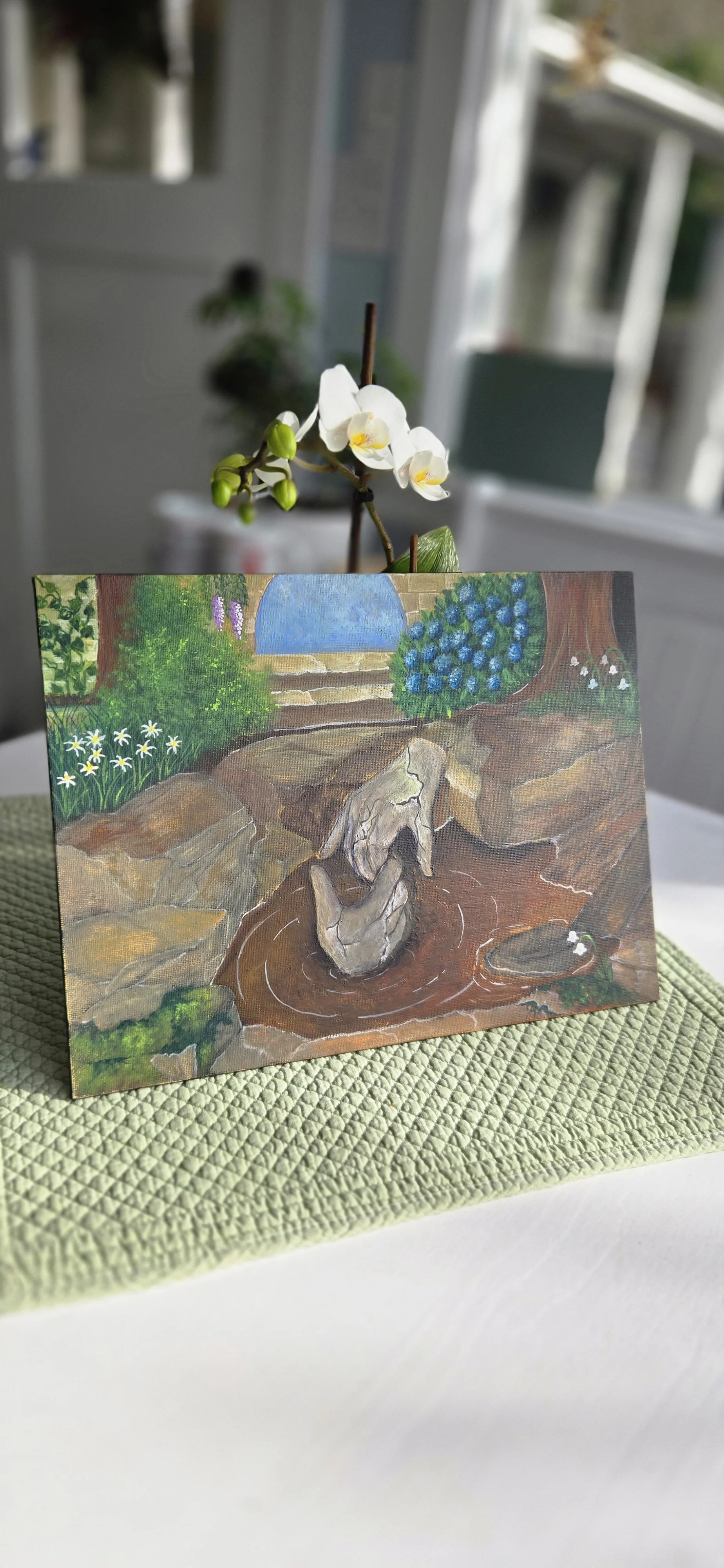 A painted landscape scene showing a small waterfall or pond with rocks, surrounded by greenery including bushes, plants, and small flowers, with a gate and a pathway in the background.