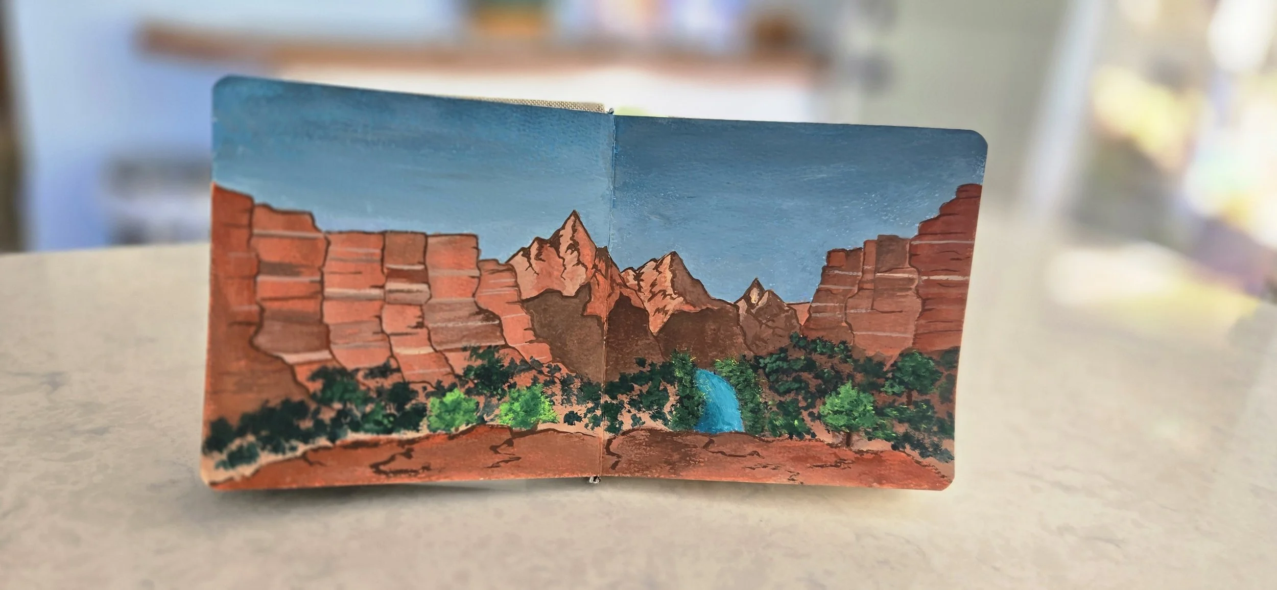 A pink and blue paper booklet featuring an illustration of a desert landscape with red rock formations, green trees, and a blue river.