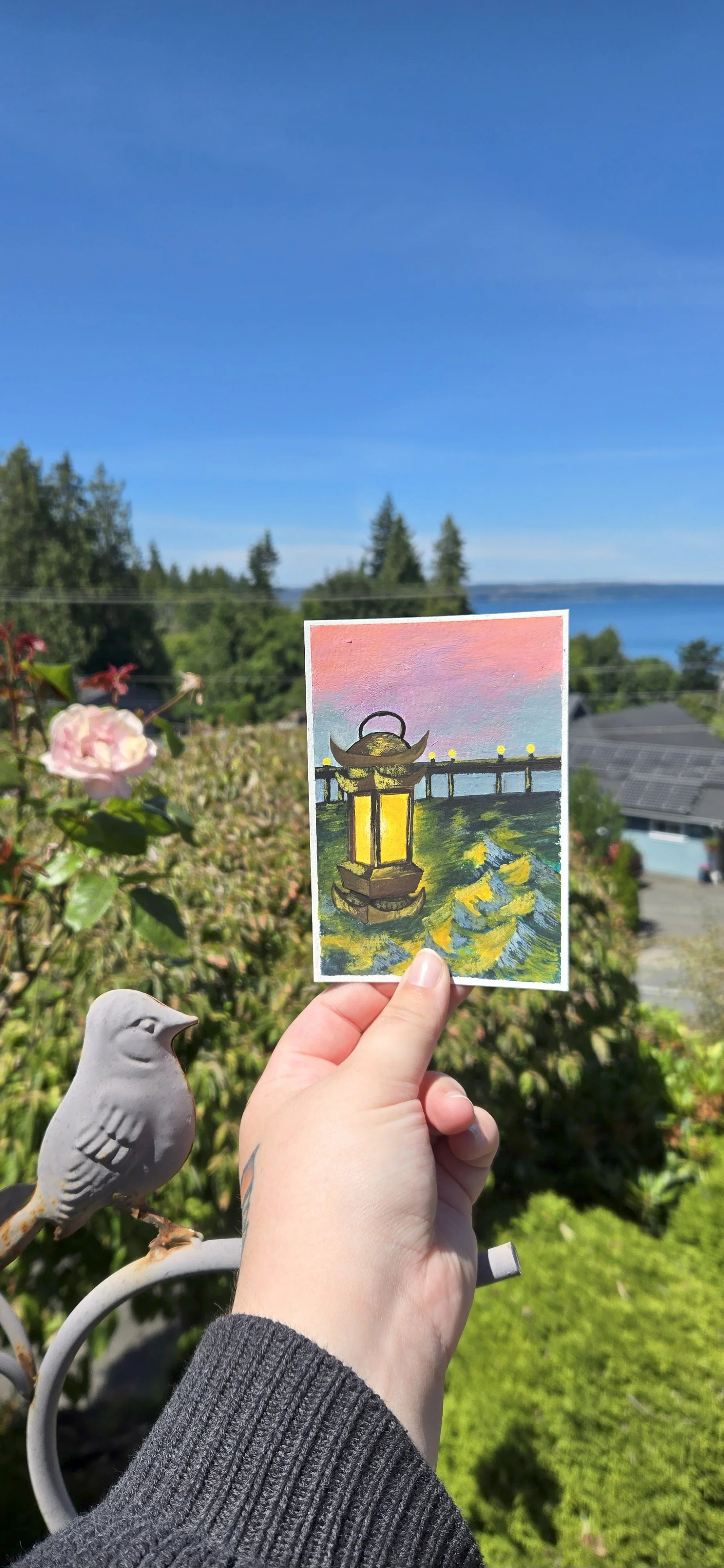 Hand holding a small painting of a traditional Japanese lantern on a pier with a colorful sky, set against a lush garden with a decorative bird sculpture, houses, trees, and a body of water in the background.
