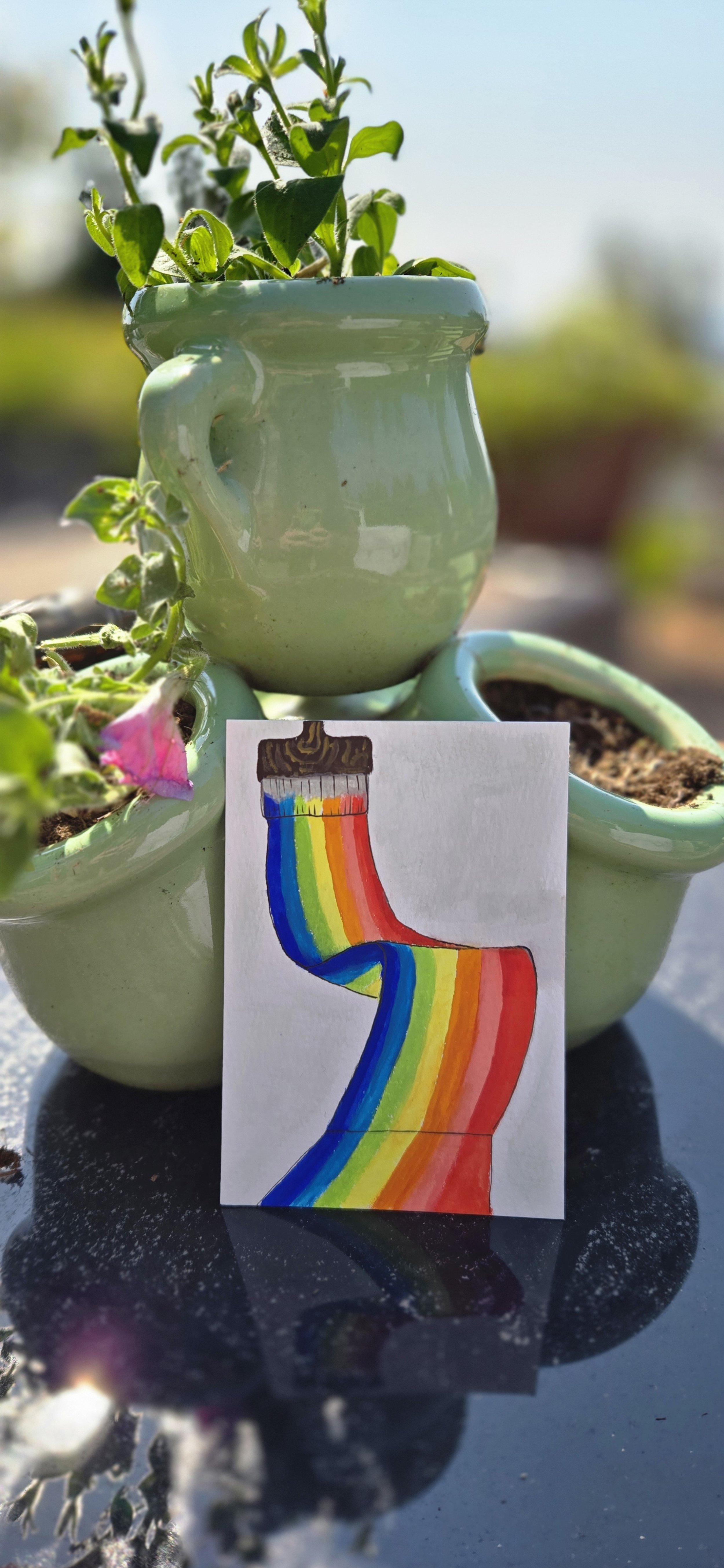 A white mug with green plants and soil in small matching pots. A colorful drawing of a rainbow-colored tube is positioned in front of the mug, reflecting on a shiny black surface.