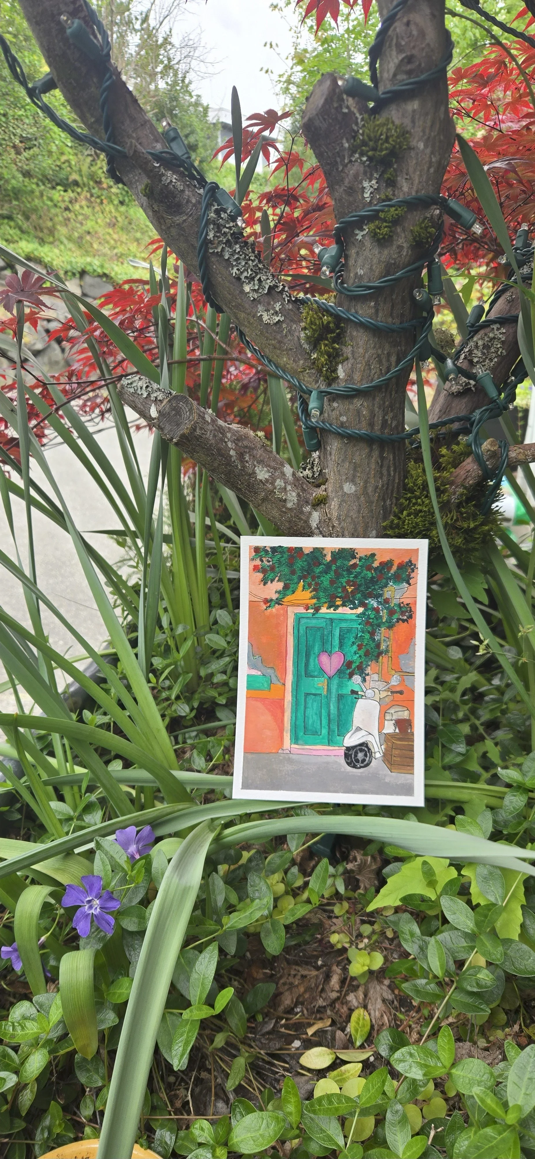 A small painting of a colorful house with a green door, a purple heart on the door, and a scooter parked outside, hangs from a tree surrounded by green plants and red foliage.