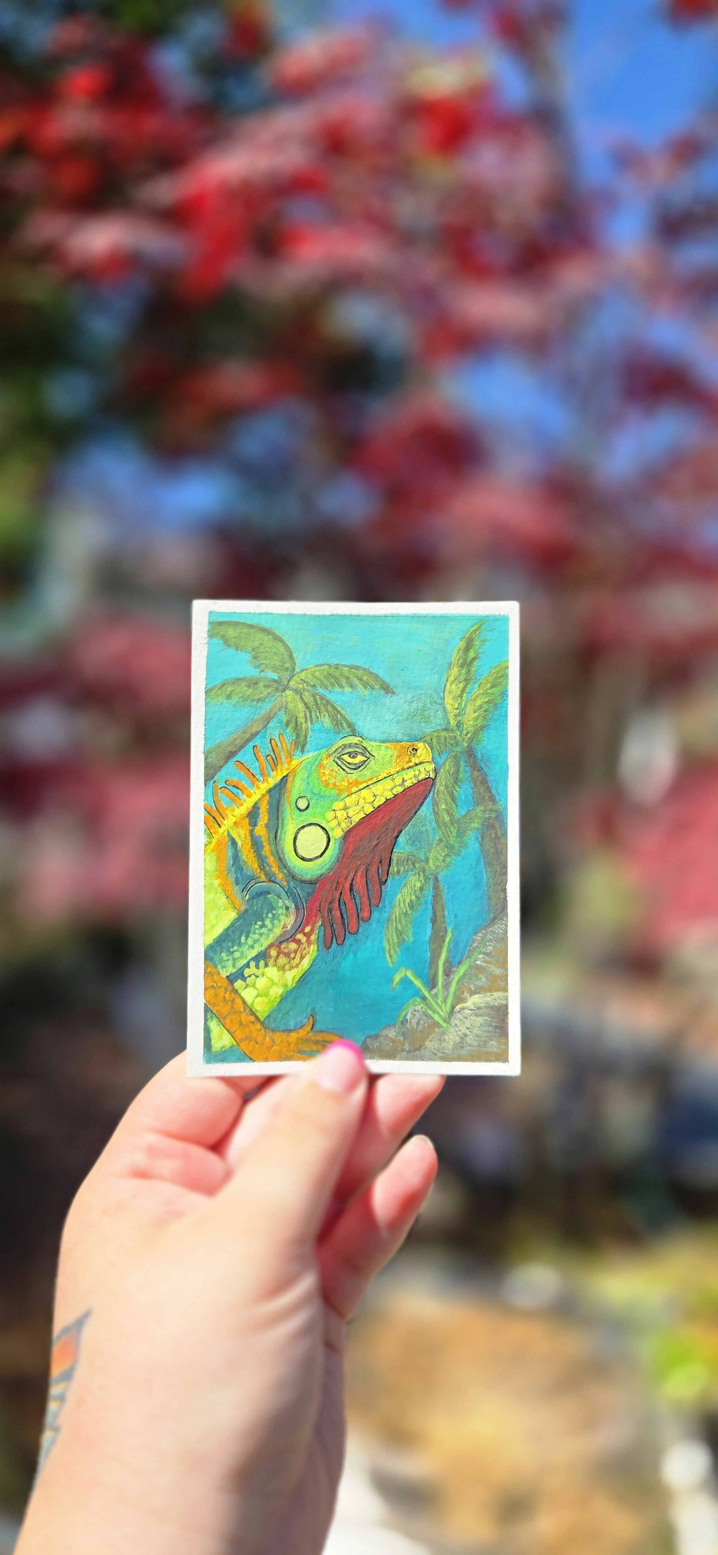 A person holding a colorful drawing of a lizard with palm trees in the background, with blurred red and blue colors in the background.