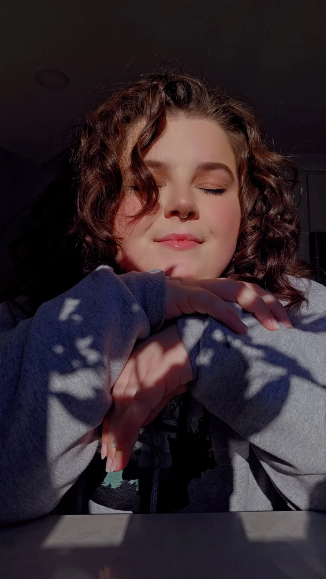 A young woman with curly brown hair and a septum piercing smiling with closed eyes, resting her chin on her hands, with sunlight casting shadows on her face and sweatshirt.
