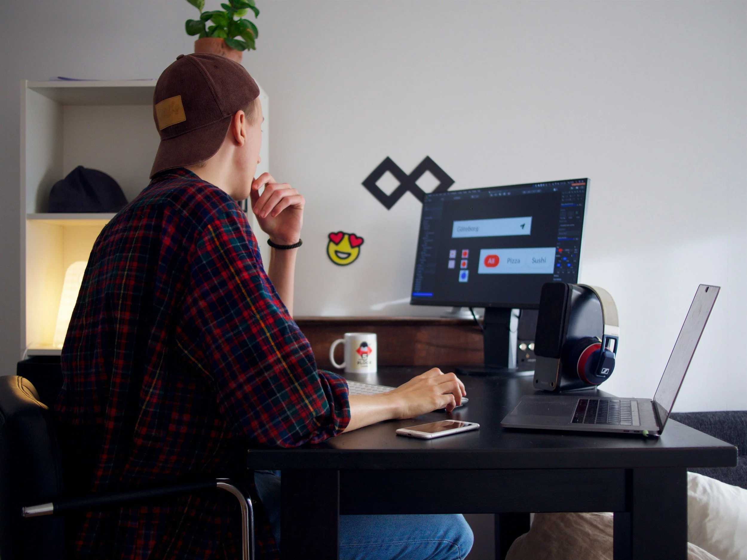 Person wearing a brown cap and plaid shirt working at a desk with a computer monitor and laptop. The monitor displays a graphic design application. A smartphone and headphones are on the desk, with a small plant and a shelf in the background.