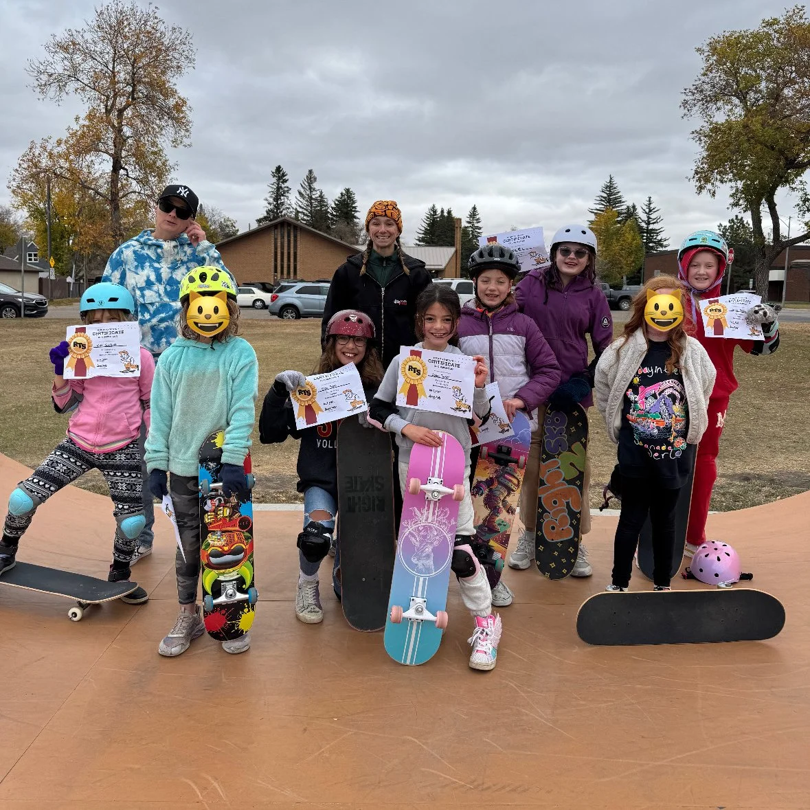 Group of children at a Right To Skate class with skateboards and helmets at skatepark, holding certificates, with two adults behind them, overcast sky, trees, and building in background.