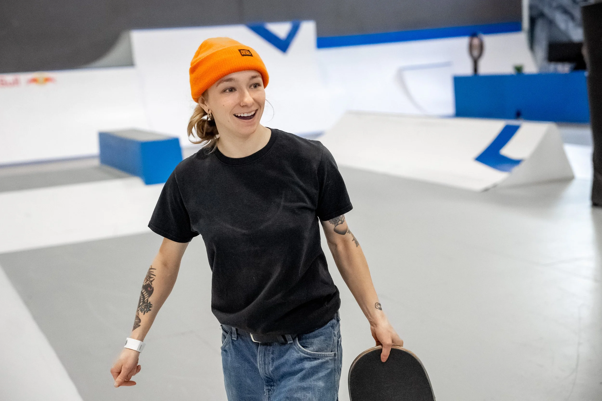 Right To Skate's Executive Director Sarah Kelly, a woman with tattoos, wearing a black t-shirt, blue jeans, and an orange beanie, smiling and holding a chair in an indoor skatepark.