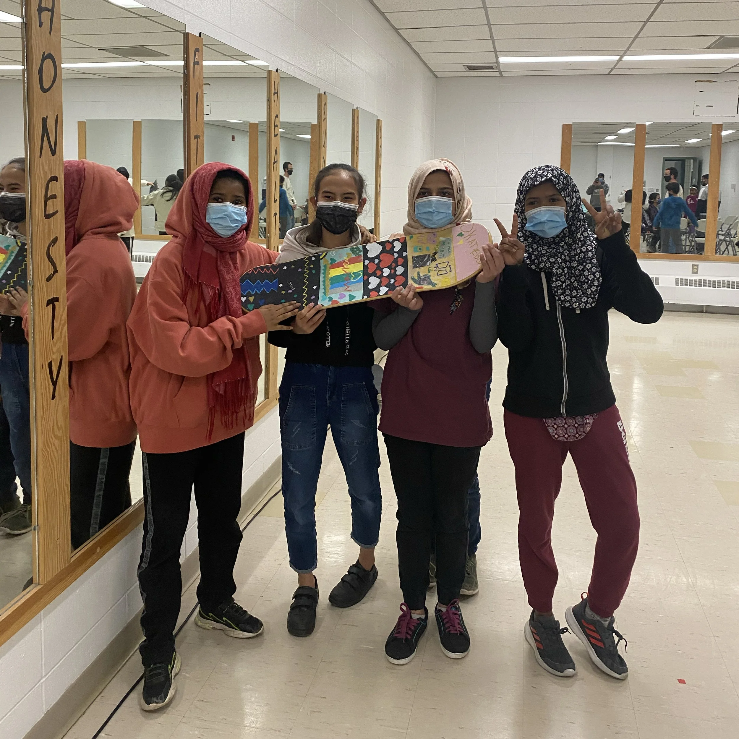 Four newcomer members of Right To Skate wearing masks standing in front of mirrors holding a colorful artwork on a skateboard.