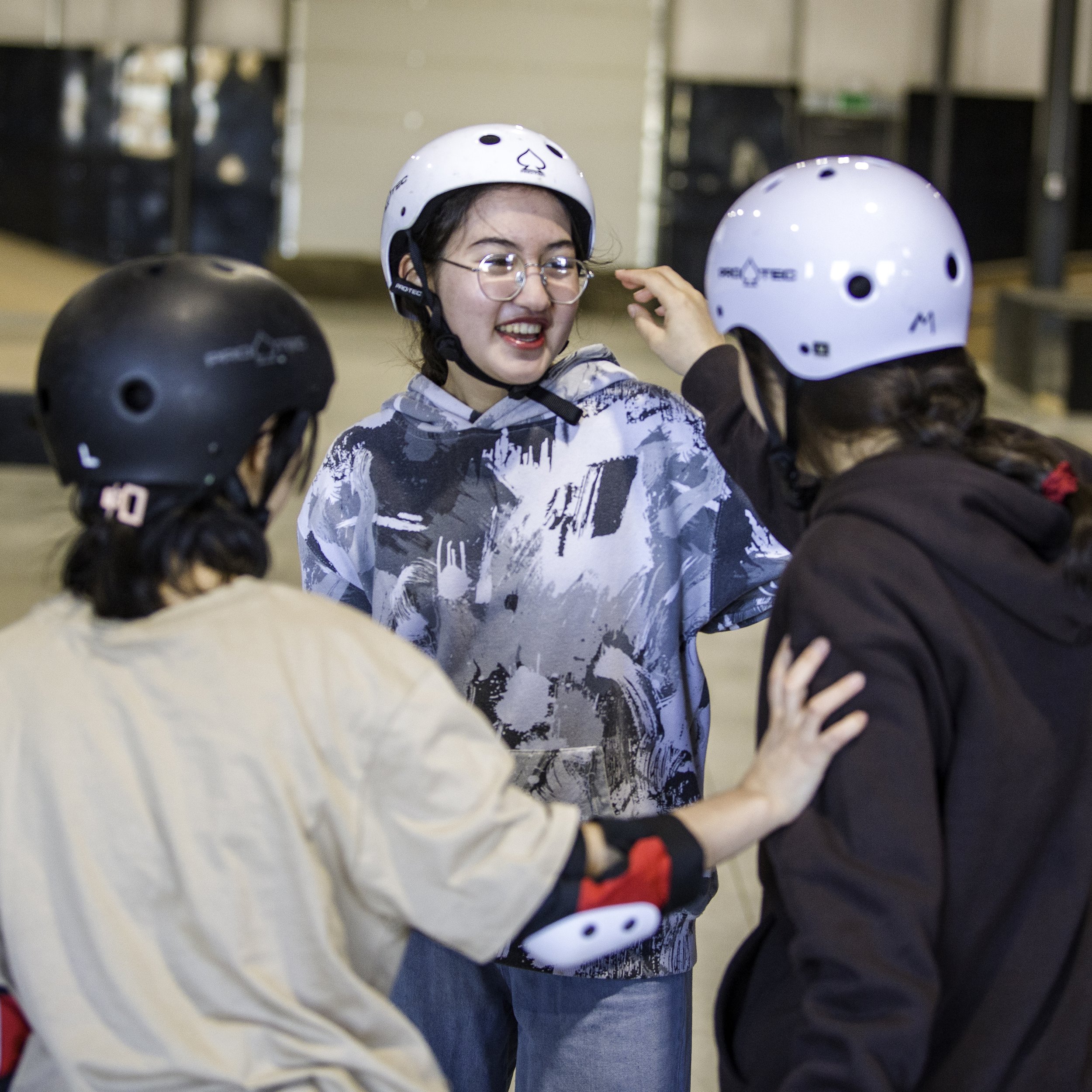 Three young girls of Right To Skate's Afghan Skate Team wearing helmets and casual clothing at an indoor skate or snowboard rink, engaged in conversation and showing friendly interactions.