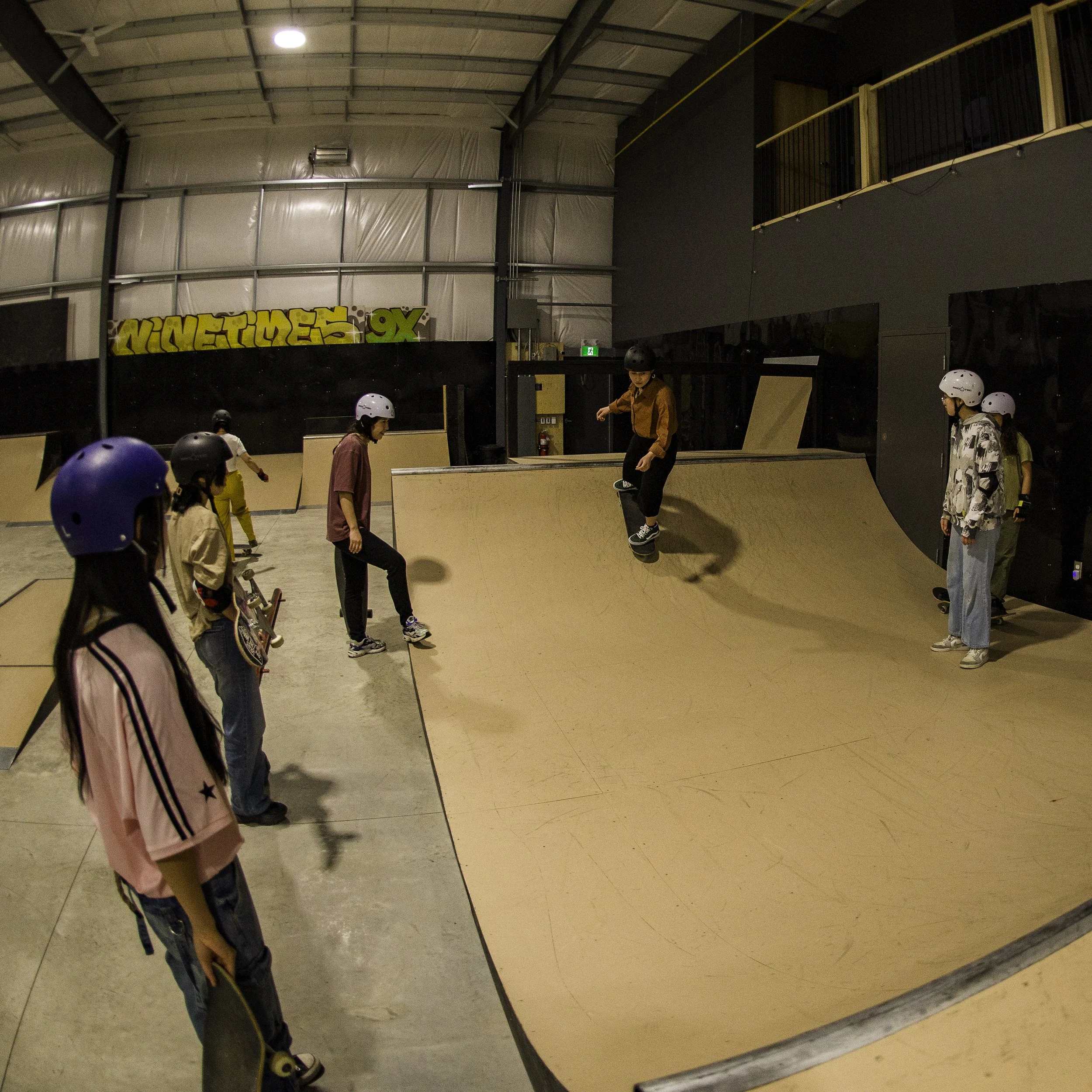 Right To Skate's Afghan Skate Team at an indoor skatepark, some wearing helmets, watching skateboarders perform tricks on a ramp.