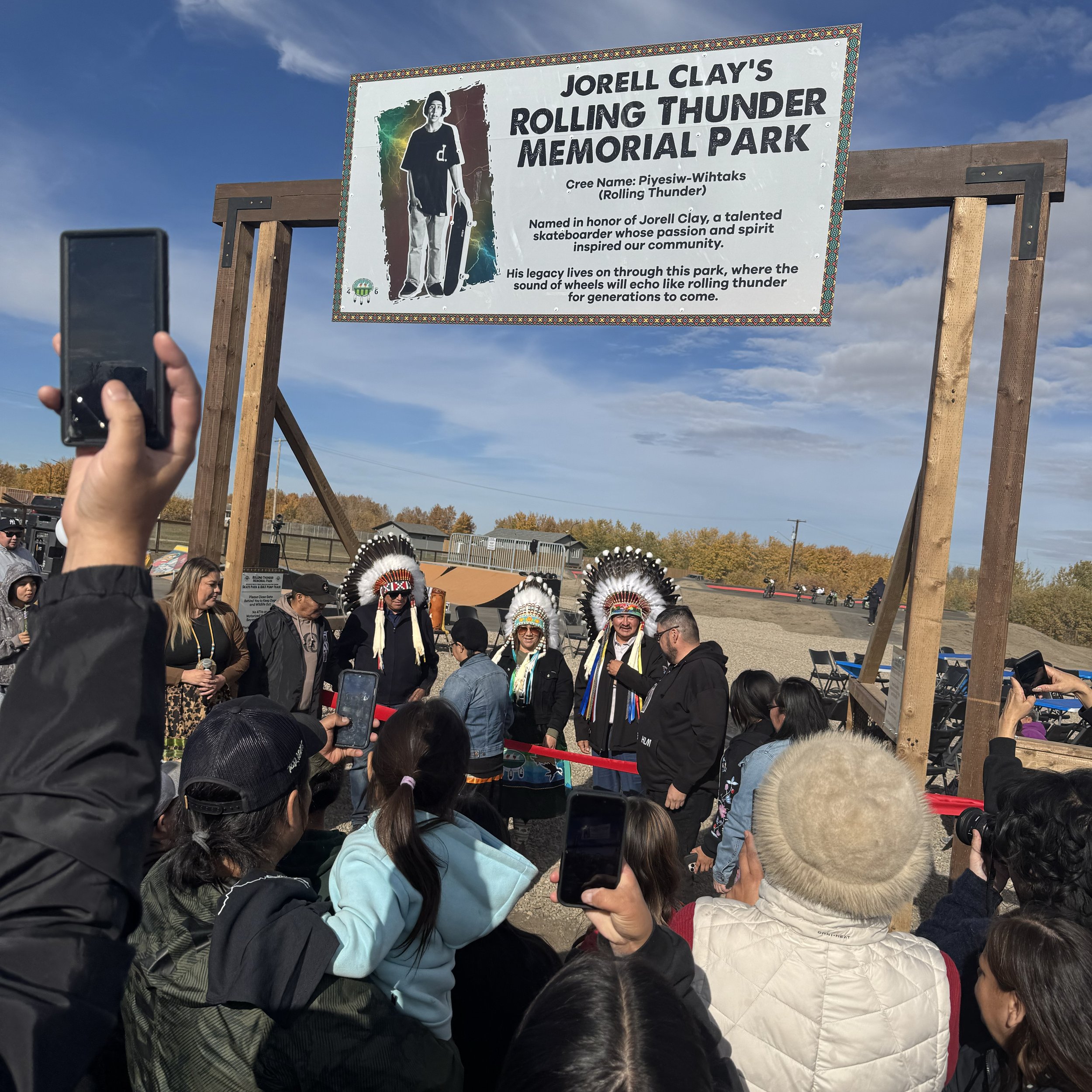 A crowd gathered at Jorell Clay's Rolling Thunder Memorial Park, with some people wearing Native American headdresses, watching and taking photos of a group of speakers or performers under a sign honoring the skateboarder Pyesiw-Wihtaks (Rolling Thun