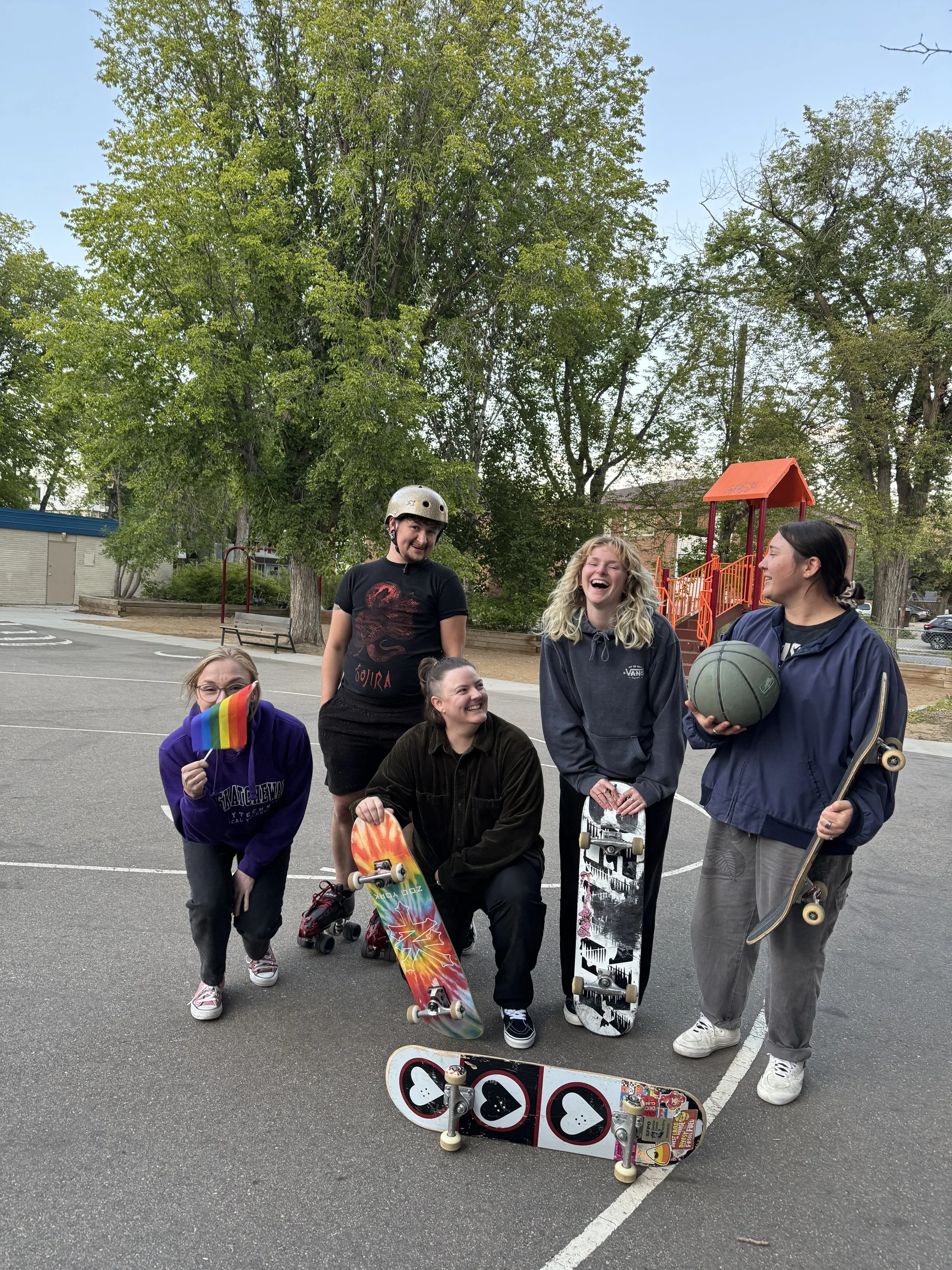 Five queer Right To Skate members with skateboards in a parking lot with trees and a playground in the background, smiling and laughing.