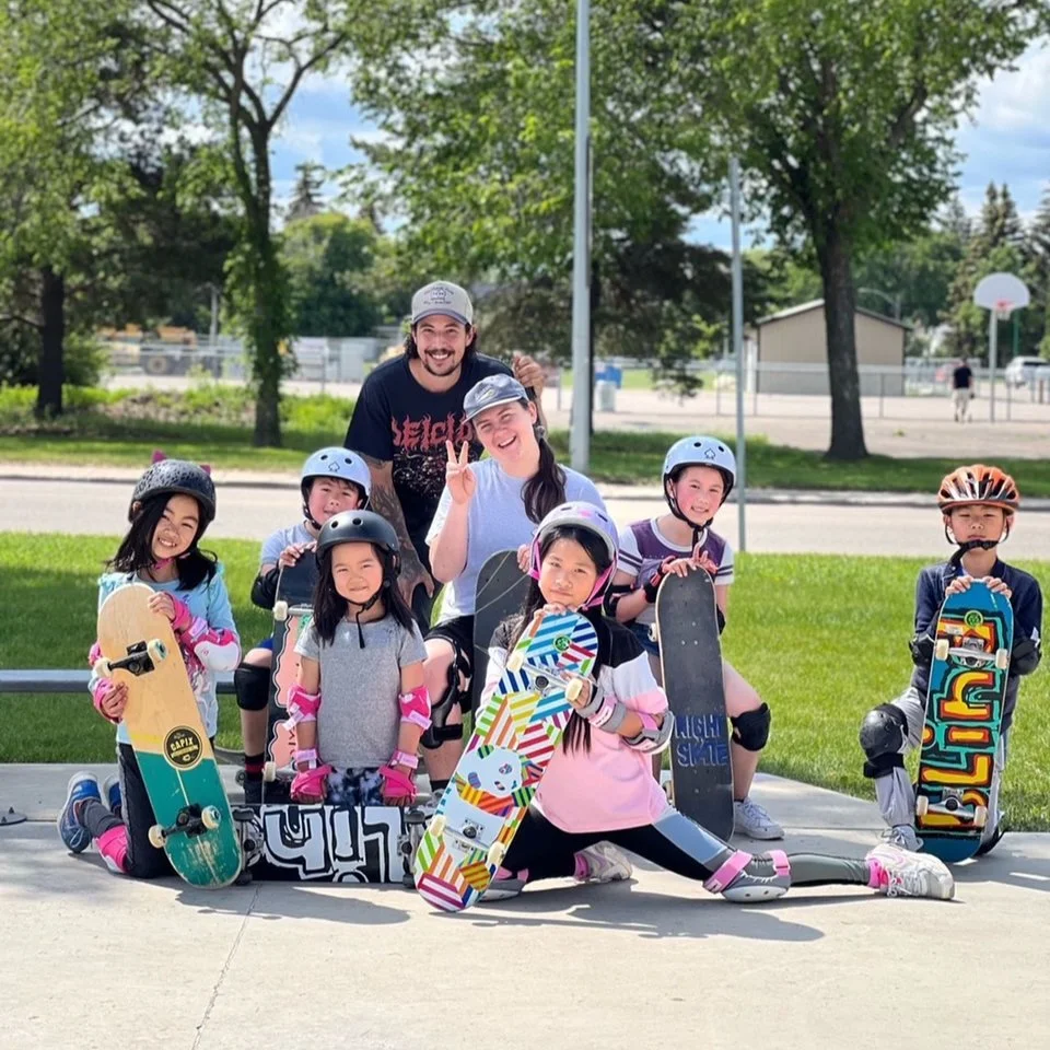 Group of kids at a Right To Skate lesson with skateboards and helmets posing outdoors at a park, smiling and giving peace signs, with two adults behind them.