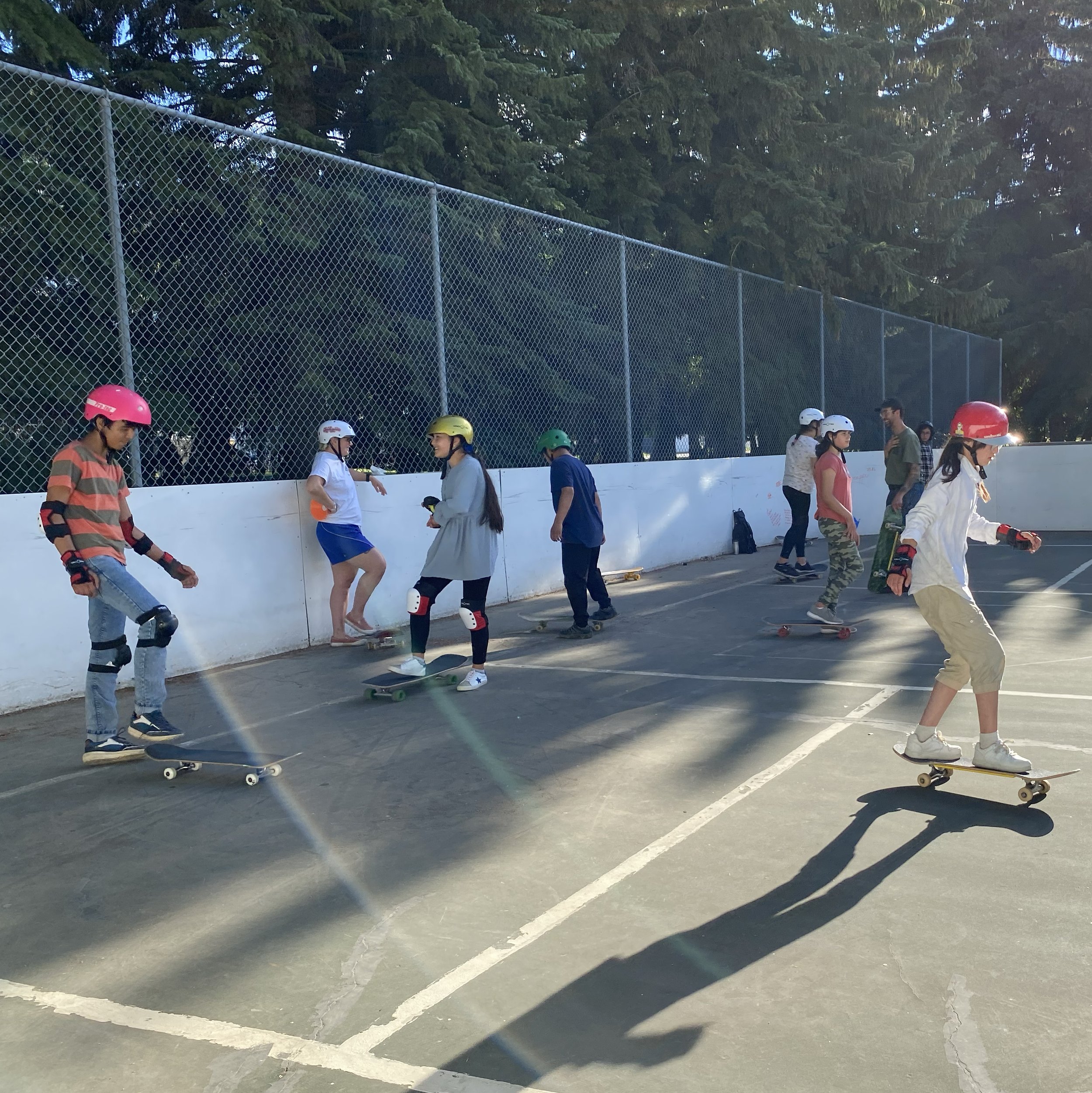 Children and teenagers in a Right To Skate class skateboarding together at an outdoor skate park surrounded by a chain-link fence and tall trees, some wearing helmets and protective gear.