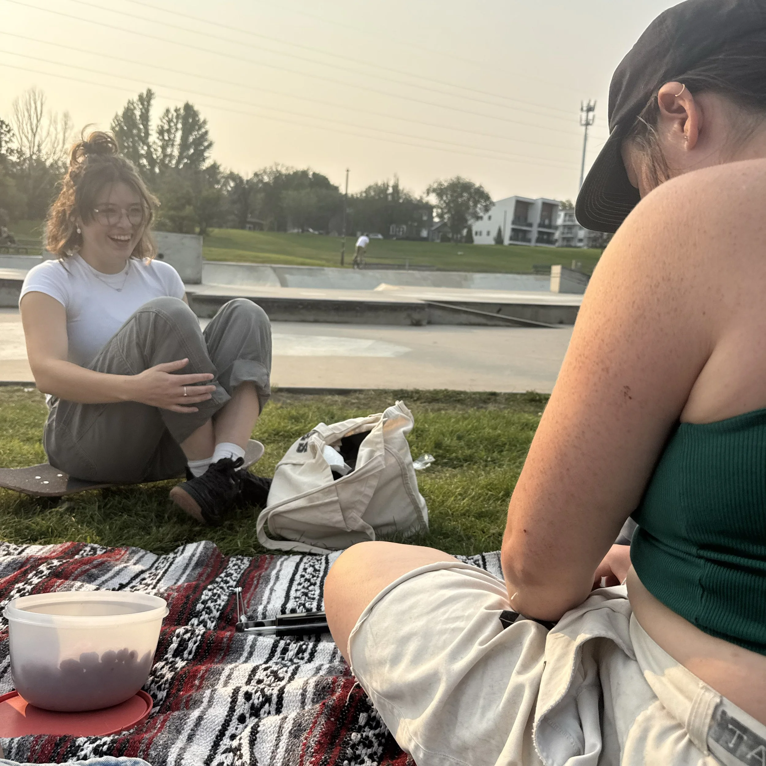 Two women sitting outdoors on a blanket, one smiling and the other with her back to the camera, near a skate park during sunset.