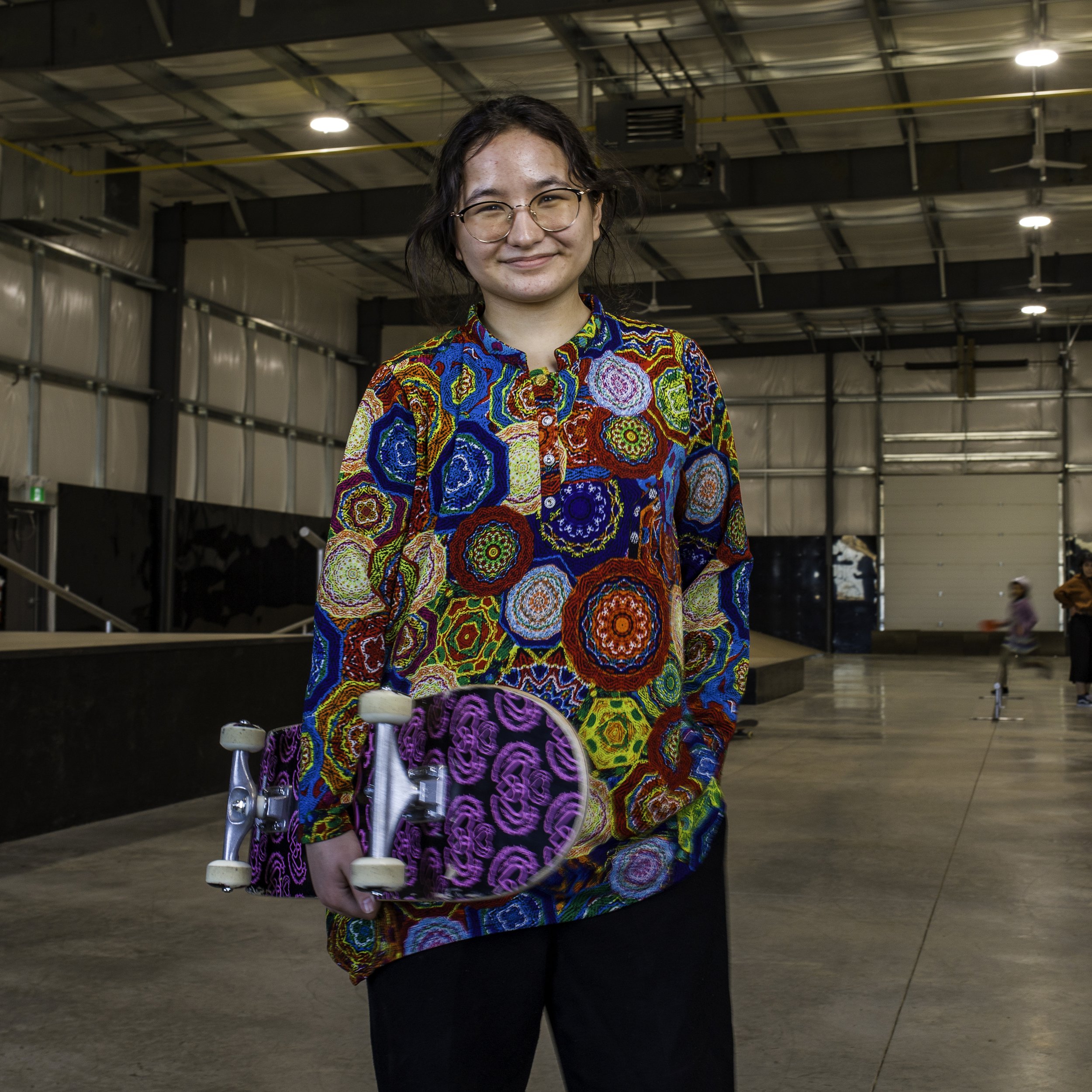 A young woman, one of Right To Skate's Afghan Team members with glasses and dark hair smiling inside a skate park. She is wearing a colourful, patterned shirt and holding a purple skateboard with a pink design.