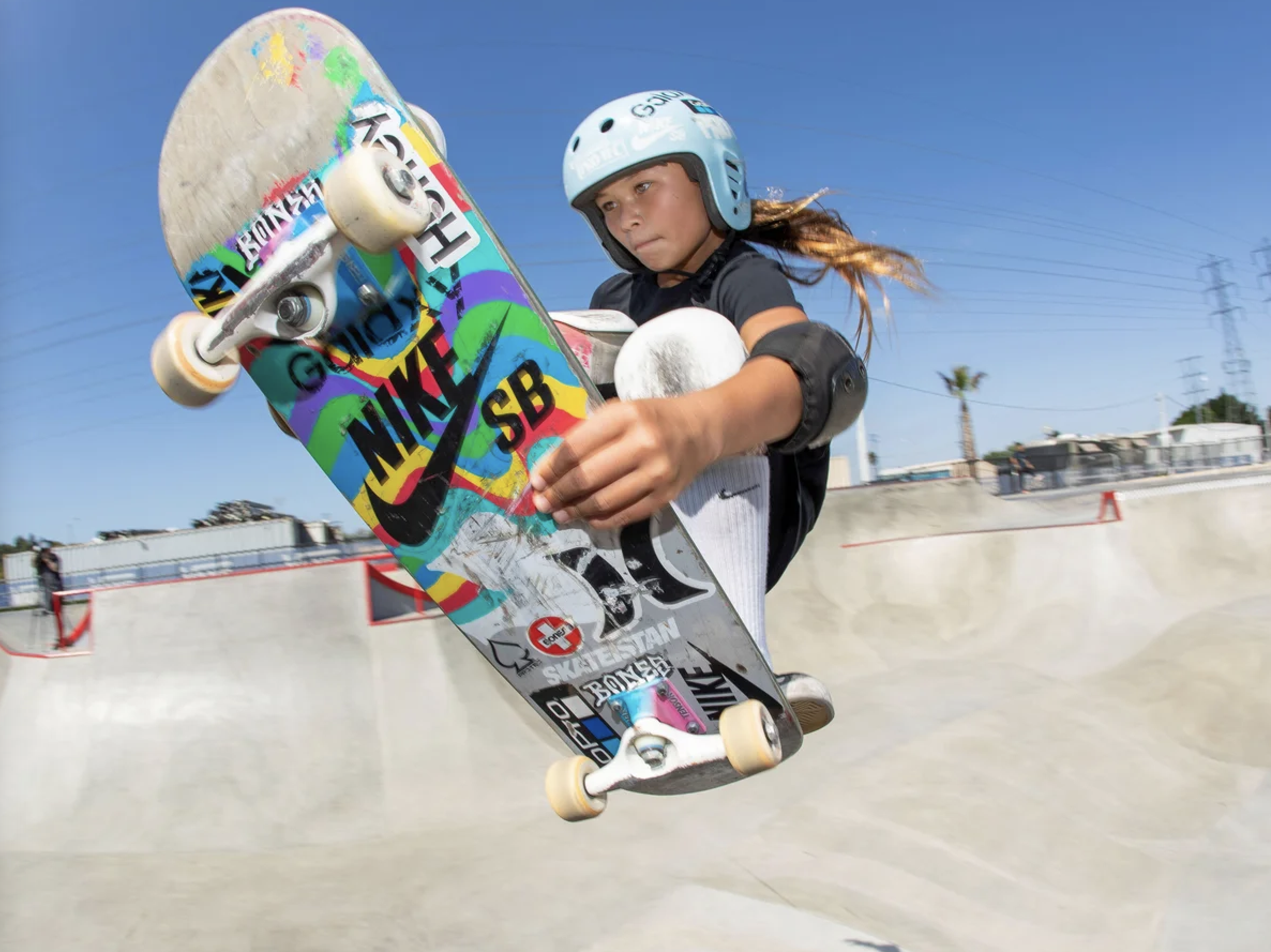 Young girl skateboarding in a skatepark on a sunny day, jumping in mid-air with a colorful skateboard.