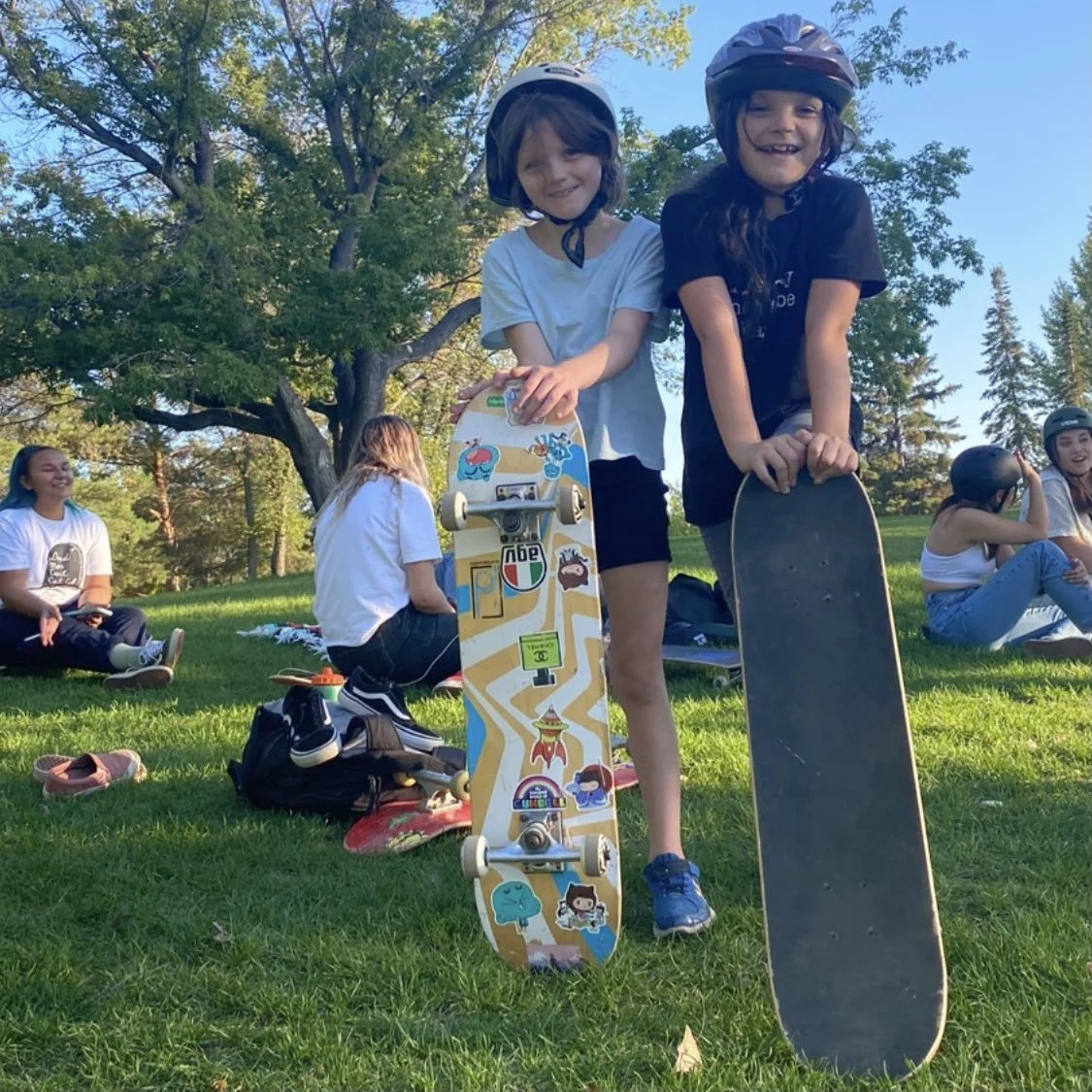 Two children at a Right To Skate class wearing helmets standing outdoors on a grassy area, each holding a skateboard, with other people sitting on the grass in the background and trees under a clear blue sky.