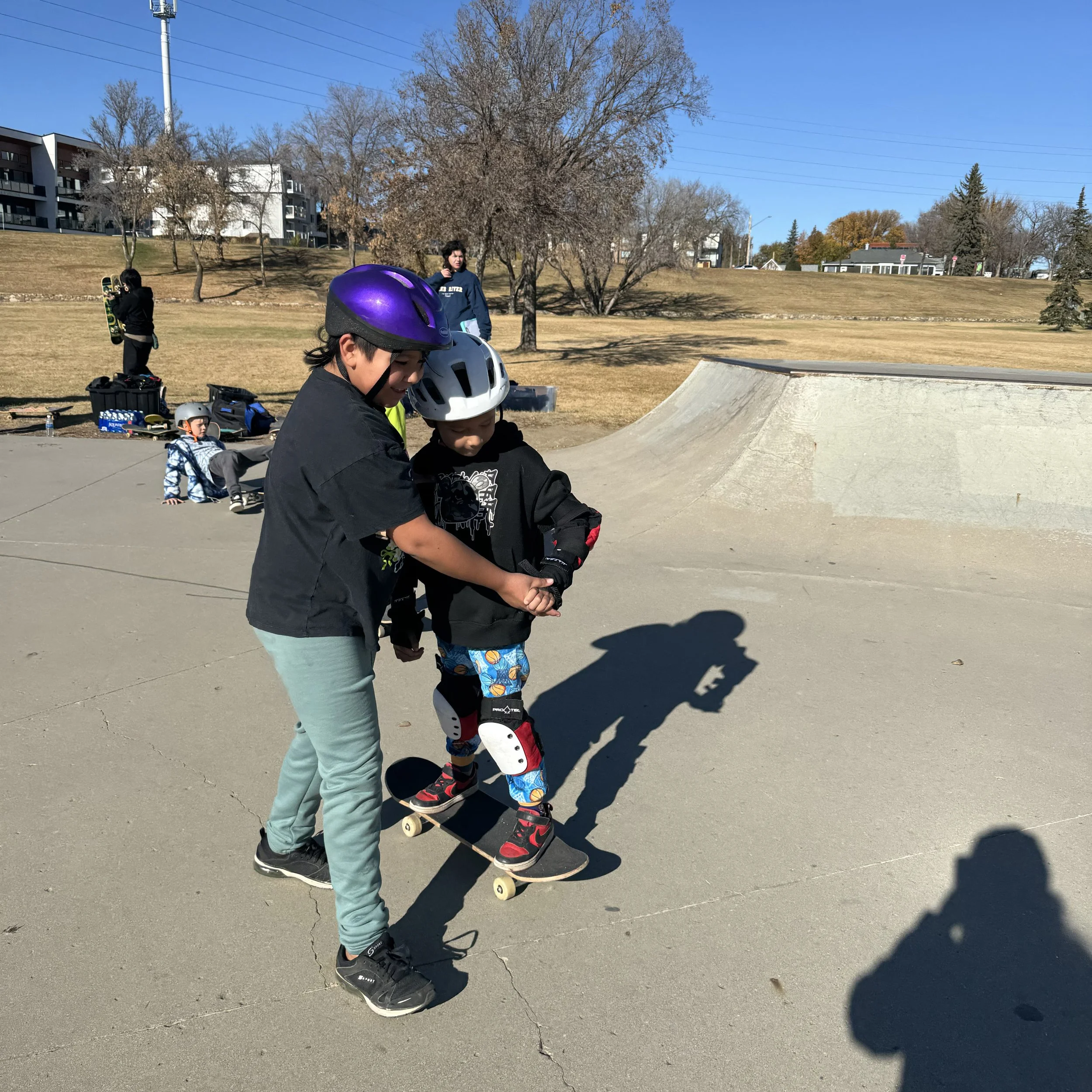 Two boys at a Right To Skate class wearing helmets and skateboards at a skate park, with one helping the other learn to skateboard, and people in the background.