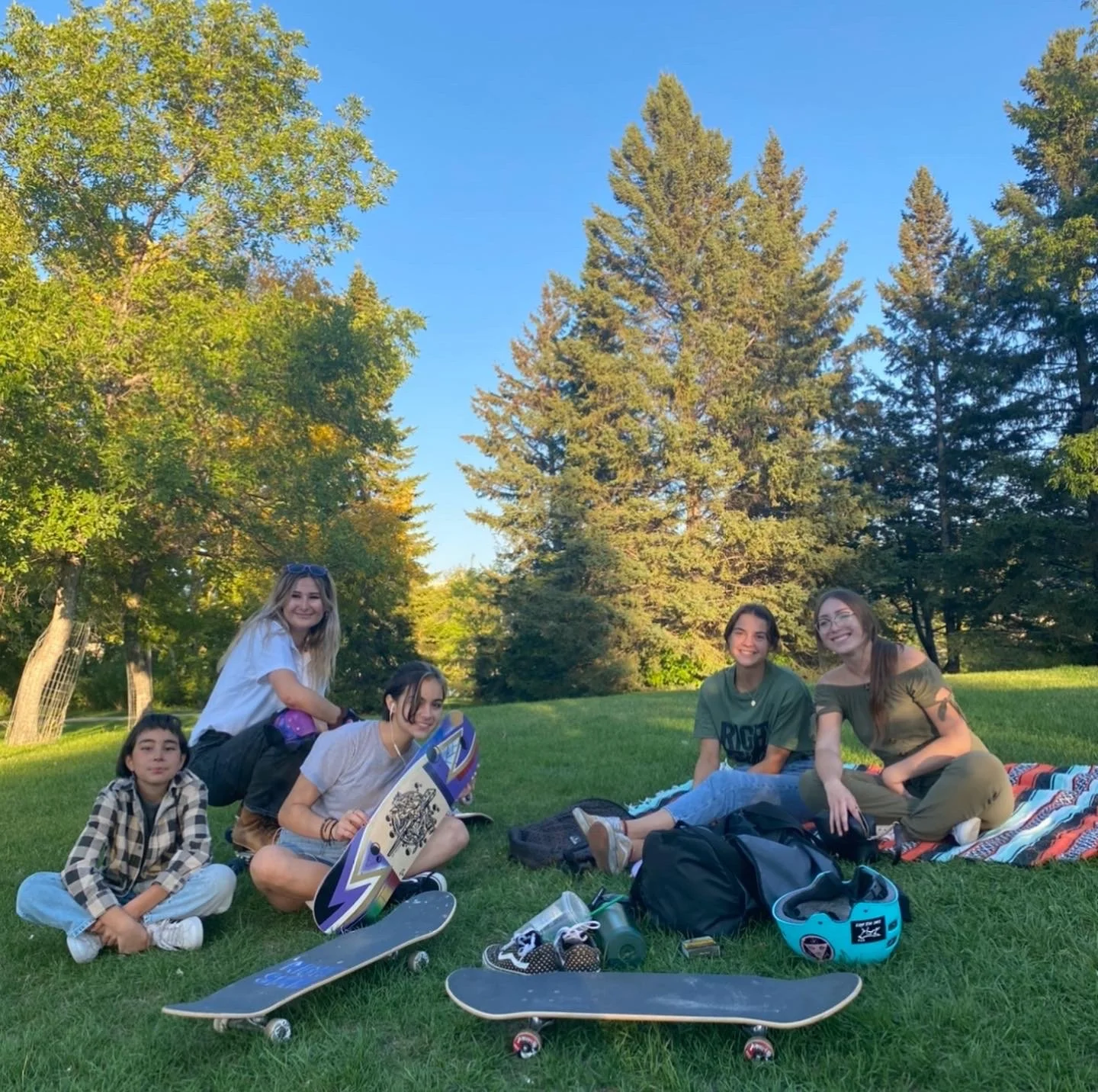 Five young women at a Right To Skate event sitting on the grass in a park with skateboard equipment around them, trees, and a bright blue sky.