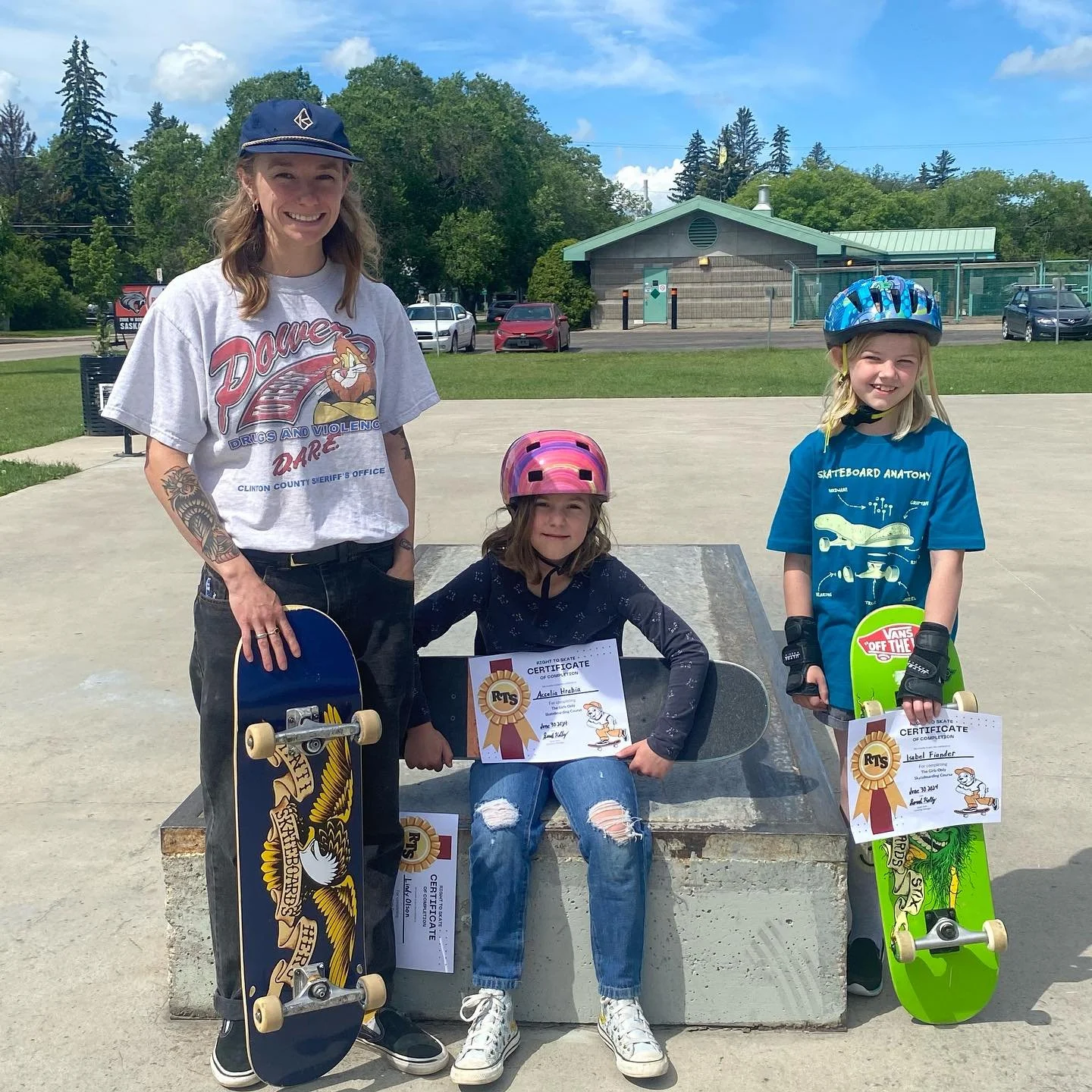 A woman and two young girls with skateboards and helmets at a skate park at a Right To Skate class. The women and one girl are holding certificates of achievement.