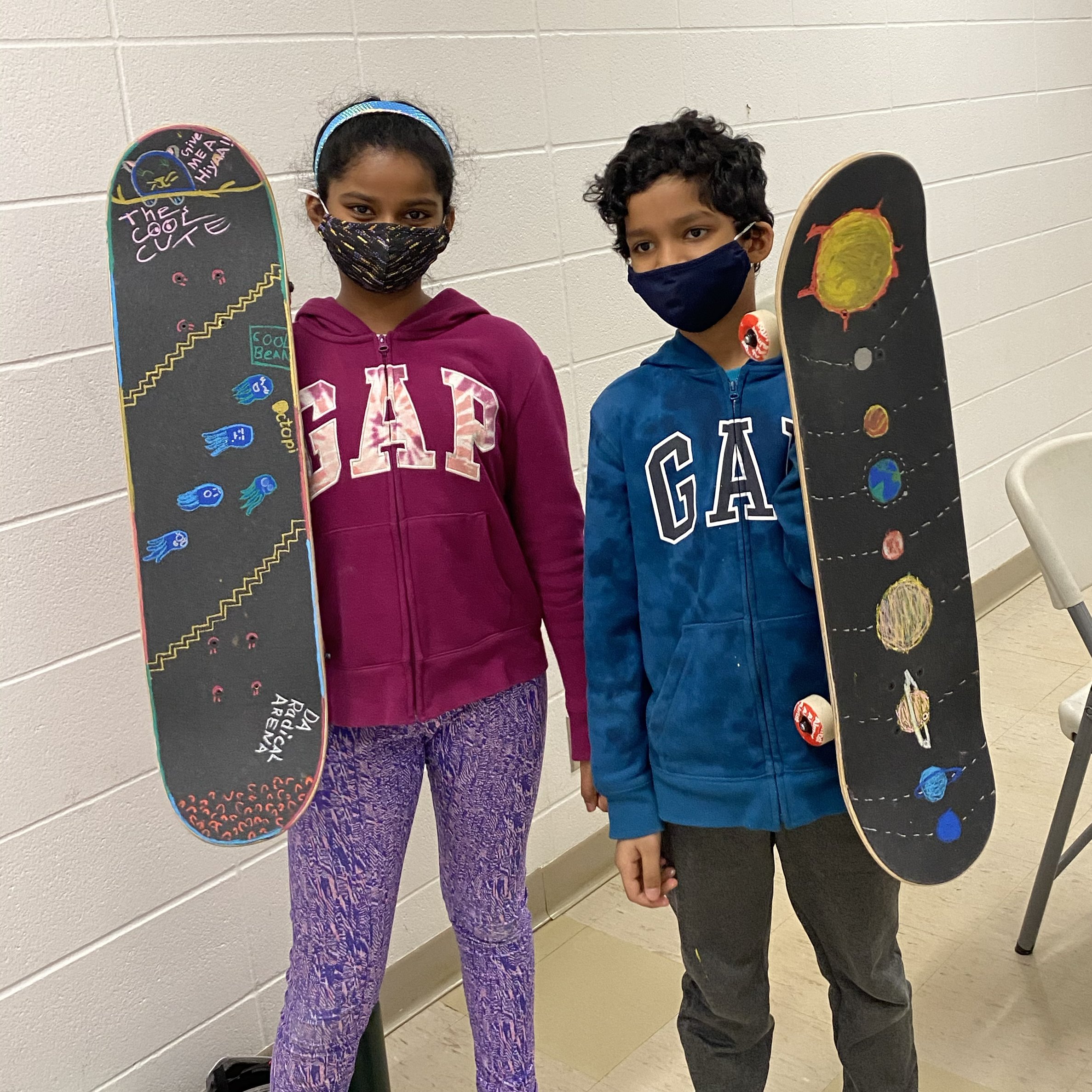 Two children in a Right To Skate class, a girl and a boy, standing indoors against a white brick wall, each holding a skateboard painted with space-themed artwork. Both are wearing face masks and casual clothes.