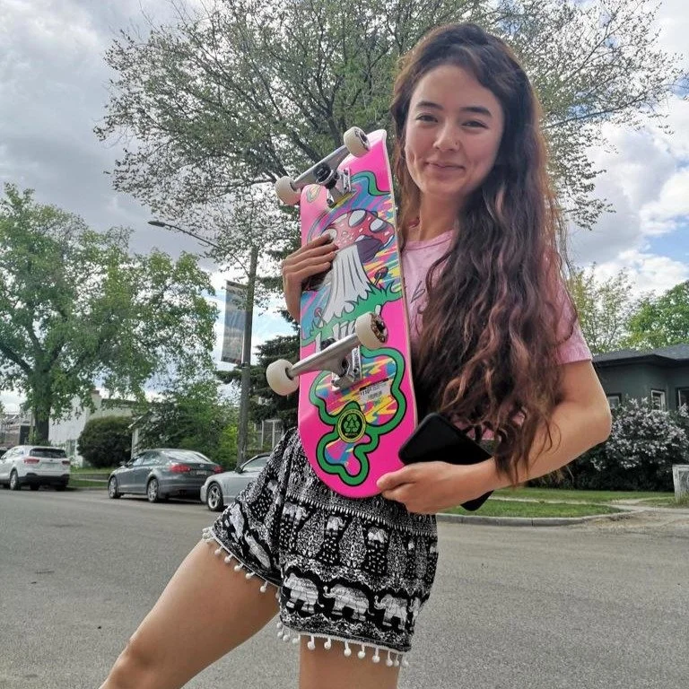 A young girl with long wavy hair smiling outdoors, holding a pink skateboard with colorful graphics, standing on a street with cars and trees in the background.