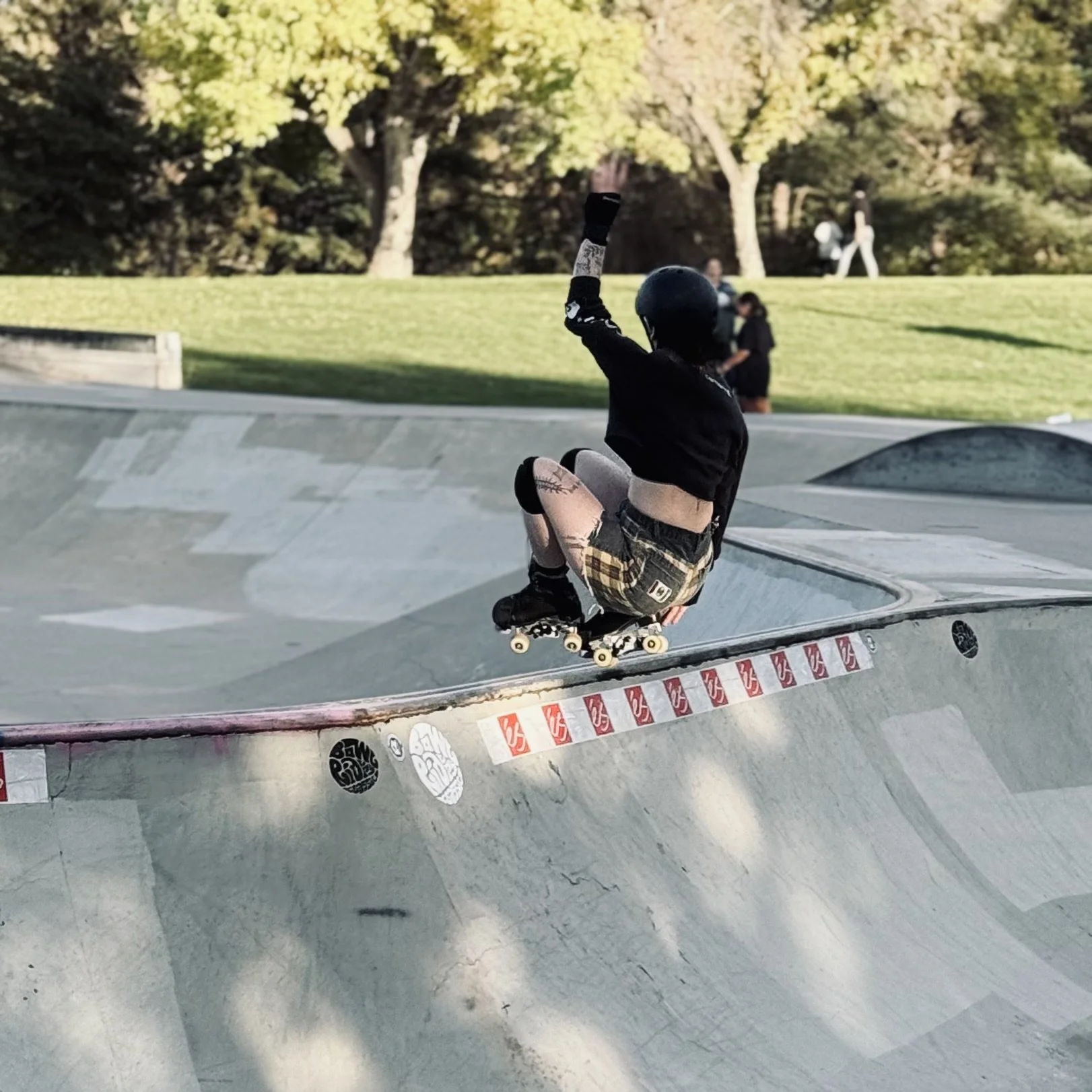 A person wearing a black helmet, black top, plaid shorts, and black skate shoes skateboarding in a concrete skate park during daytime.
