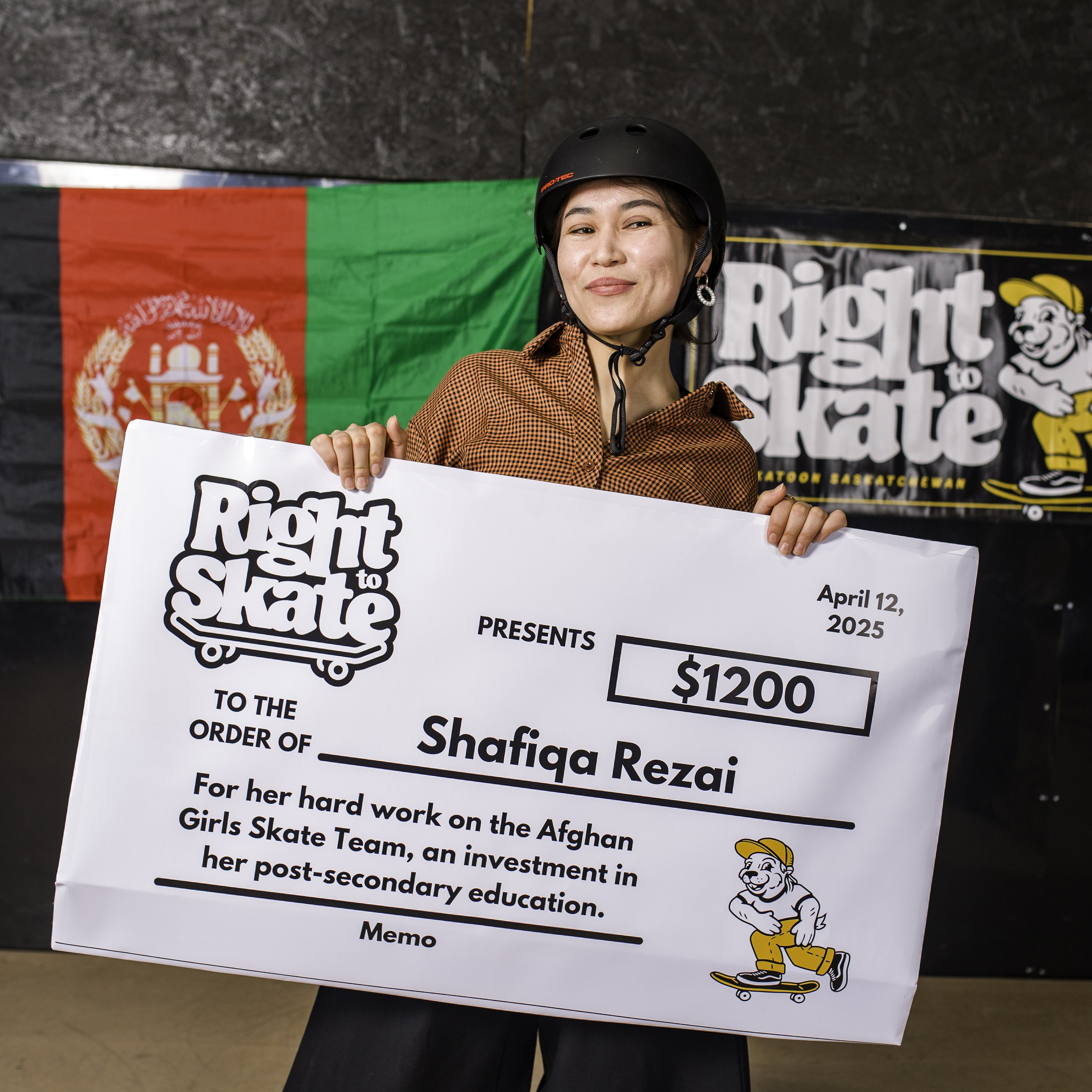 A woman wearing a black helmet and orange checkered shirt holding a large mock check for $1200 from Rights to Skate, awarded to Shafiqa Rezai for her work on the Afghan Girls Skate Team, with Afghanistan flag in the background.