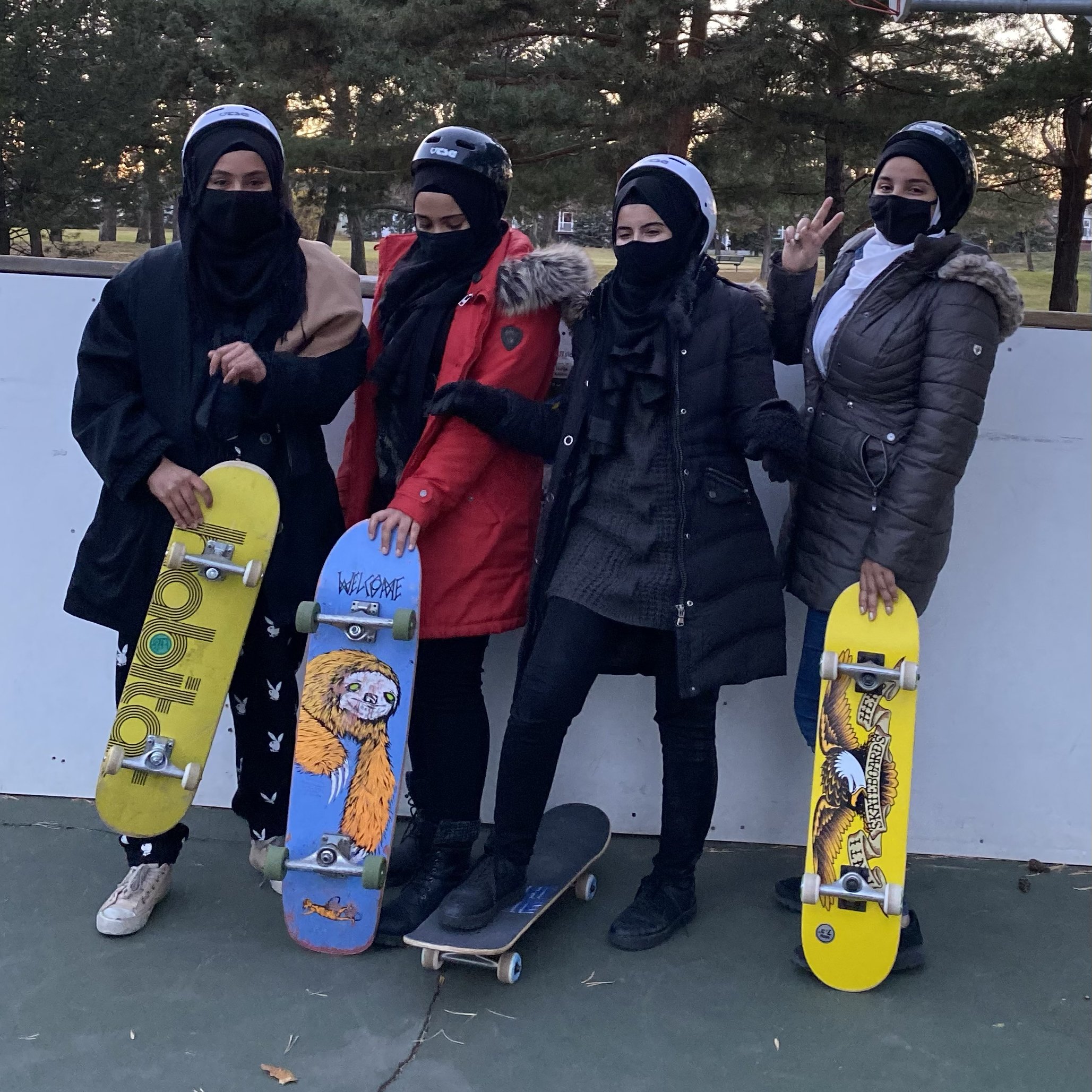 Four newcomer women in a Right To Skate class standing outdoors, holding skateboards, wearing face masks, helmets, and winter jackets, with trees in the background.