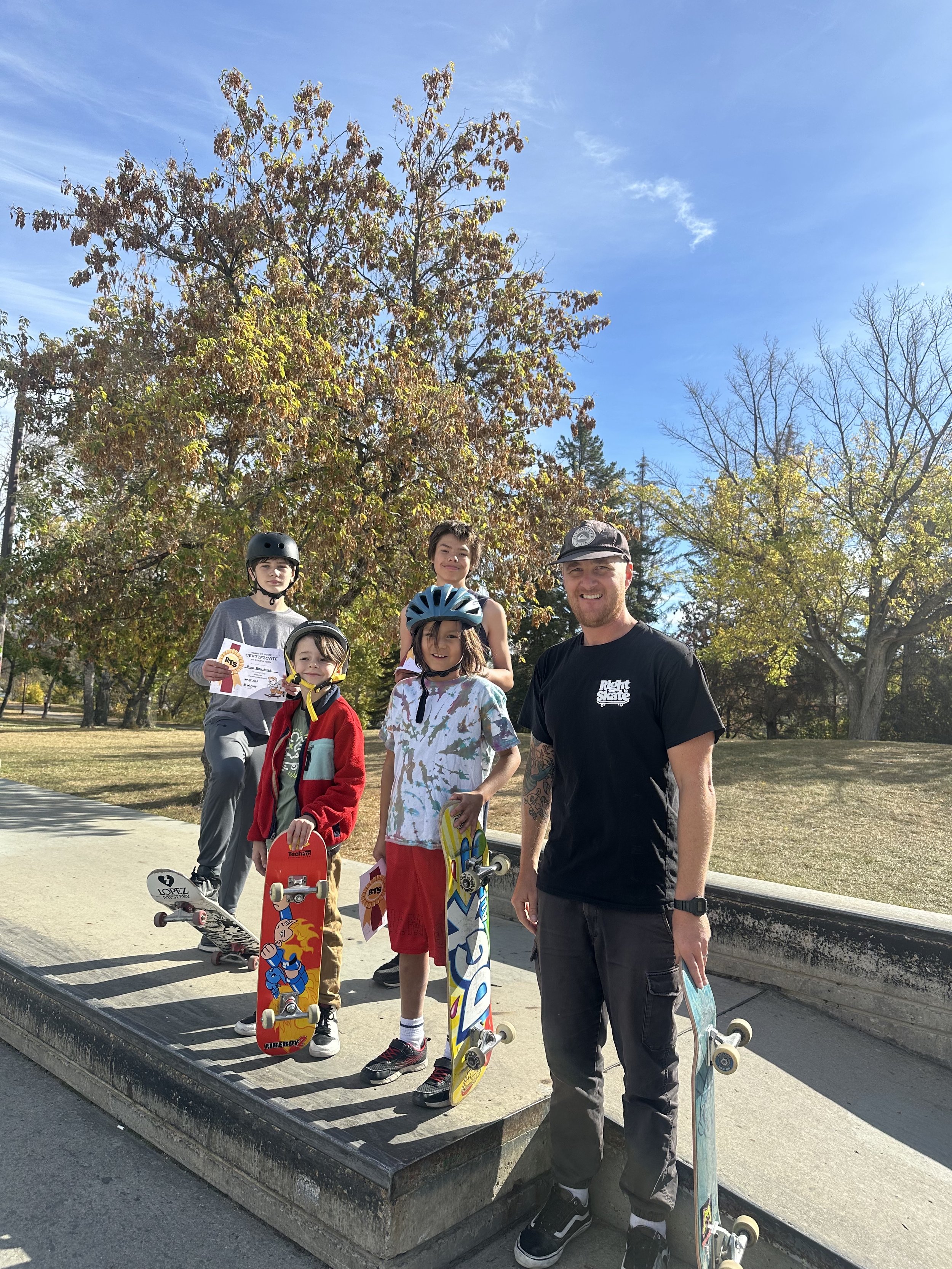 Three young girls at a skate park, two are holding skateboards and wearing helmets, and one is sitting on a concrete block holding Right To Skate certificates. The girls are smiling, and the scene is outdoors on a sunny day. 