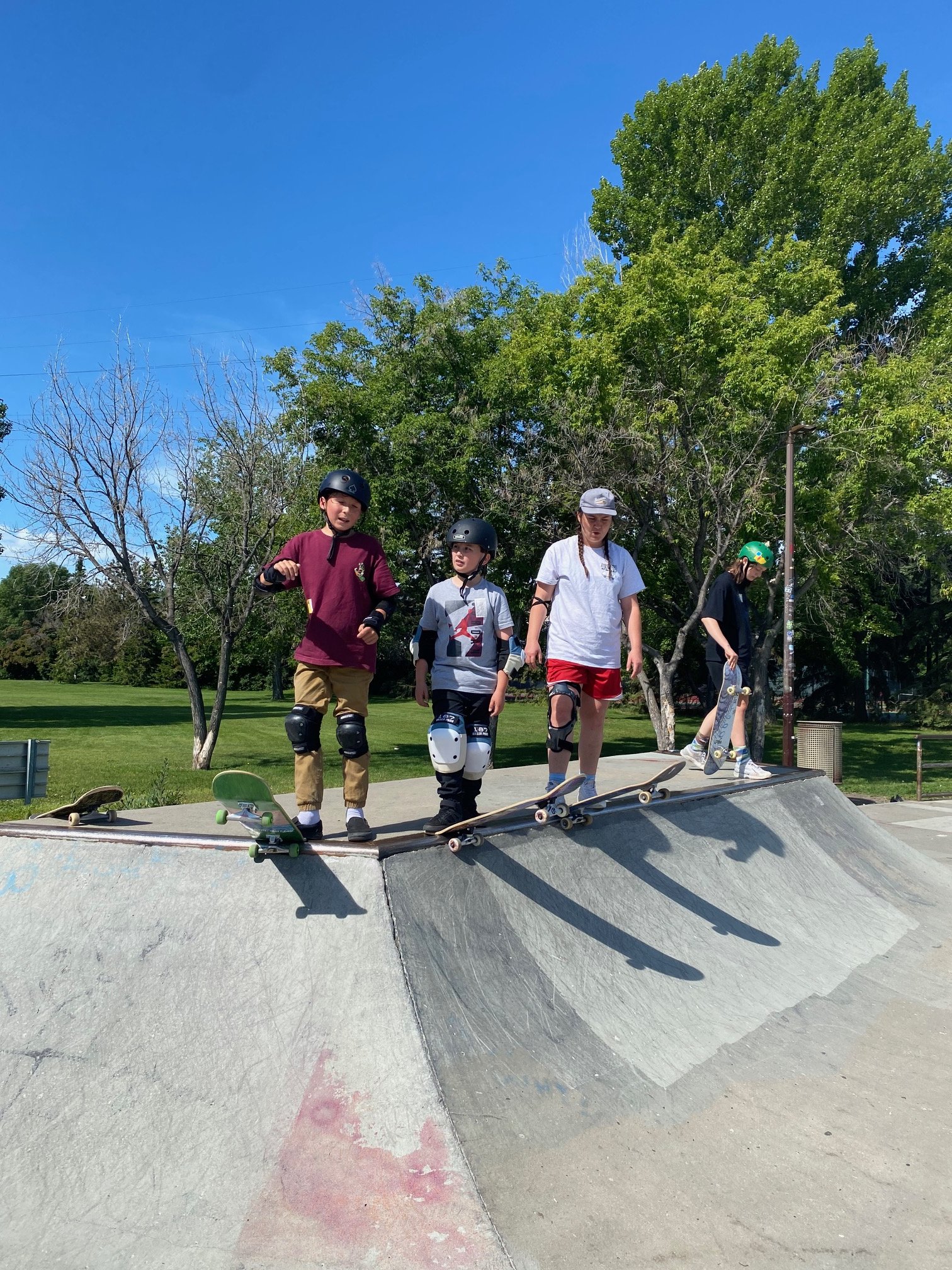 Young woman with short dark hair smiling at the camera, holding a skateboard vertically in front of her, wearing a white T-shirt with the words "Afghan Girls Skate Team Est. 2023" and a map of Afghanistan printed on it, at an outdoor skate park. 