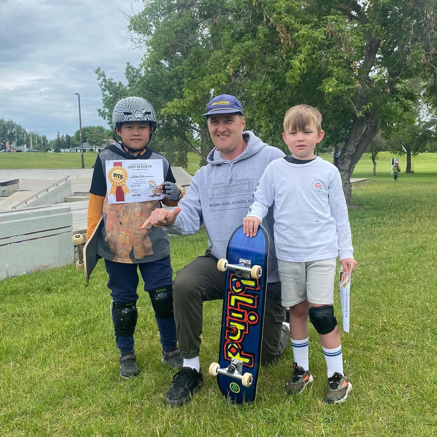 A man kneeling on the grass, holding a skateboard and giving a thumbs-up, with two young boys holding Right To Skate certificates and skateboards standing beside him, outdoors in a park with trees and a cloudy sky.