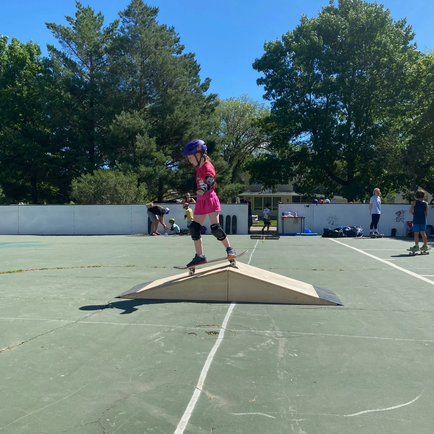 A young girl in a Right To Skate class skateboards on a small wooden ramp on an outdoor skate court during daytime, wearing a pink dress, purple helmet, and protective gear.
