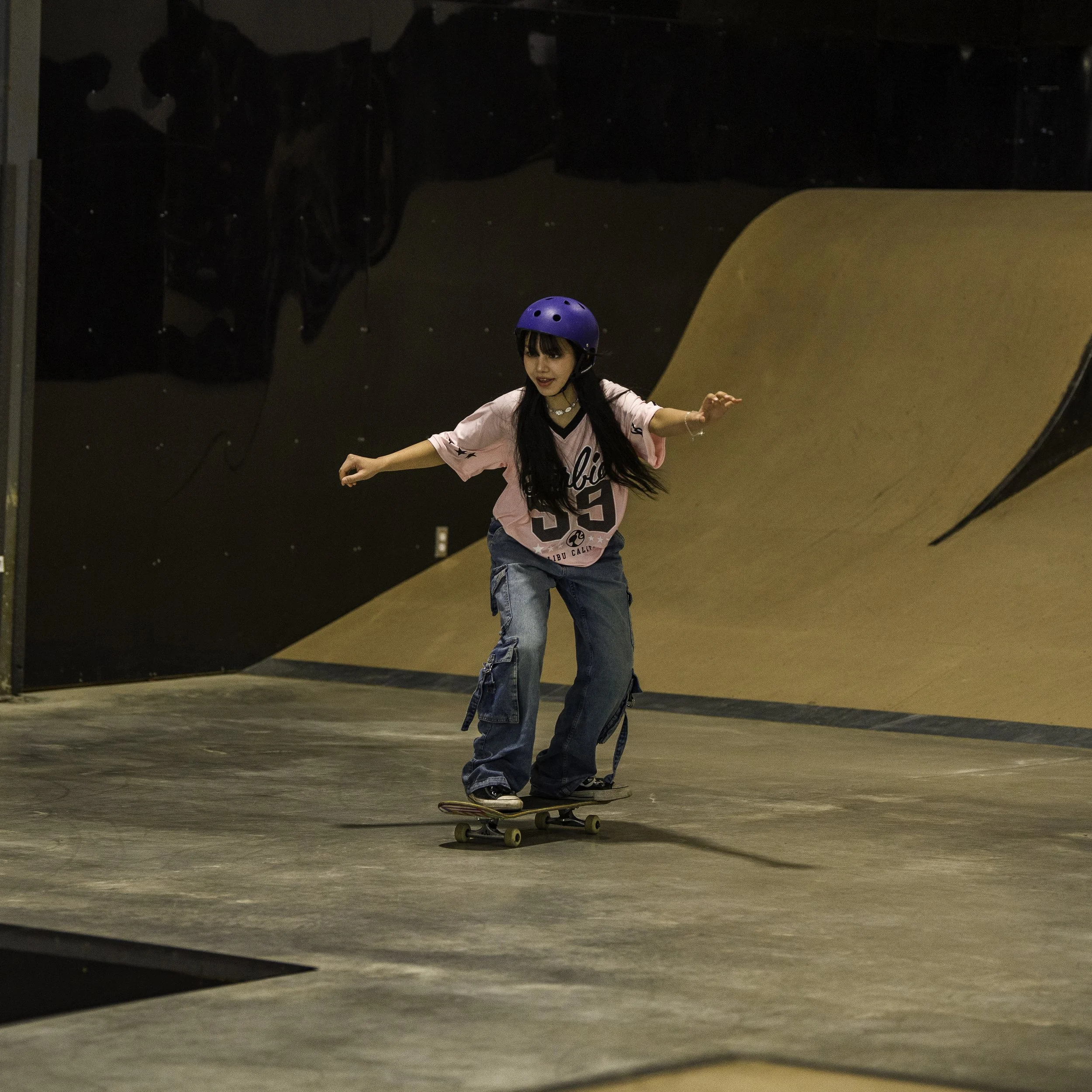 A young girl of Right To Skate's Afghan Skate Team skateboarding indoors on a ramp, wearing a blue helmet and a pink shirt.