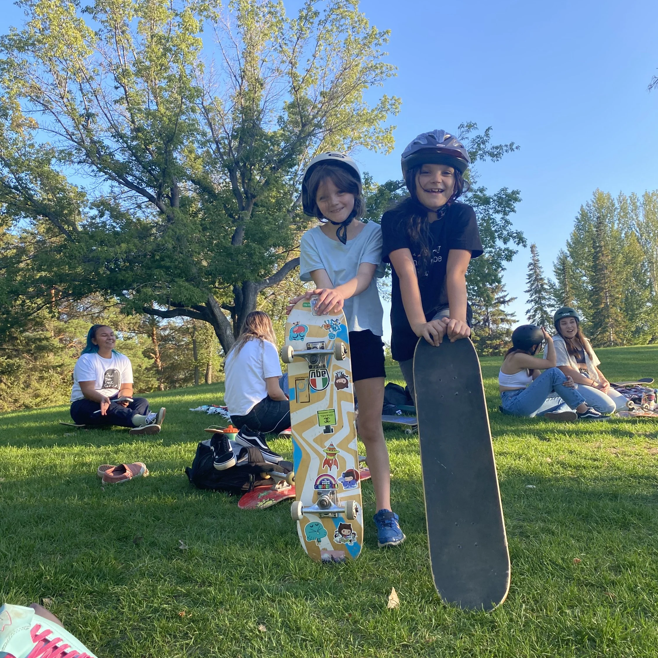 Two young girls at a Right To Skate class smiling and holding skateboards, sitting on a grassy field with a group of friends in the background, all wearing helmets, under a large tree on a sunny day.