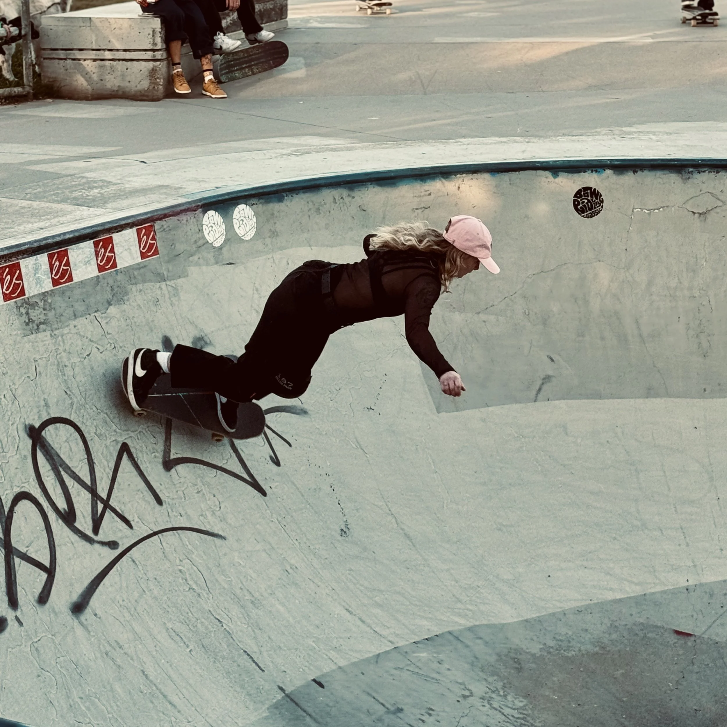 A woman in black clothing and a pink cap skateboarding inside a concrete skate bowl at a skate park.