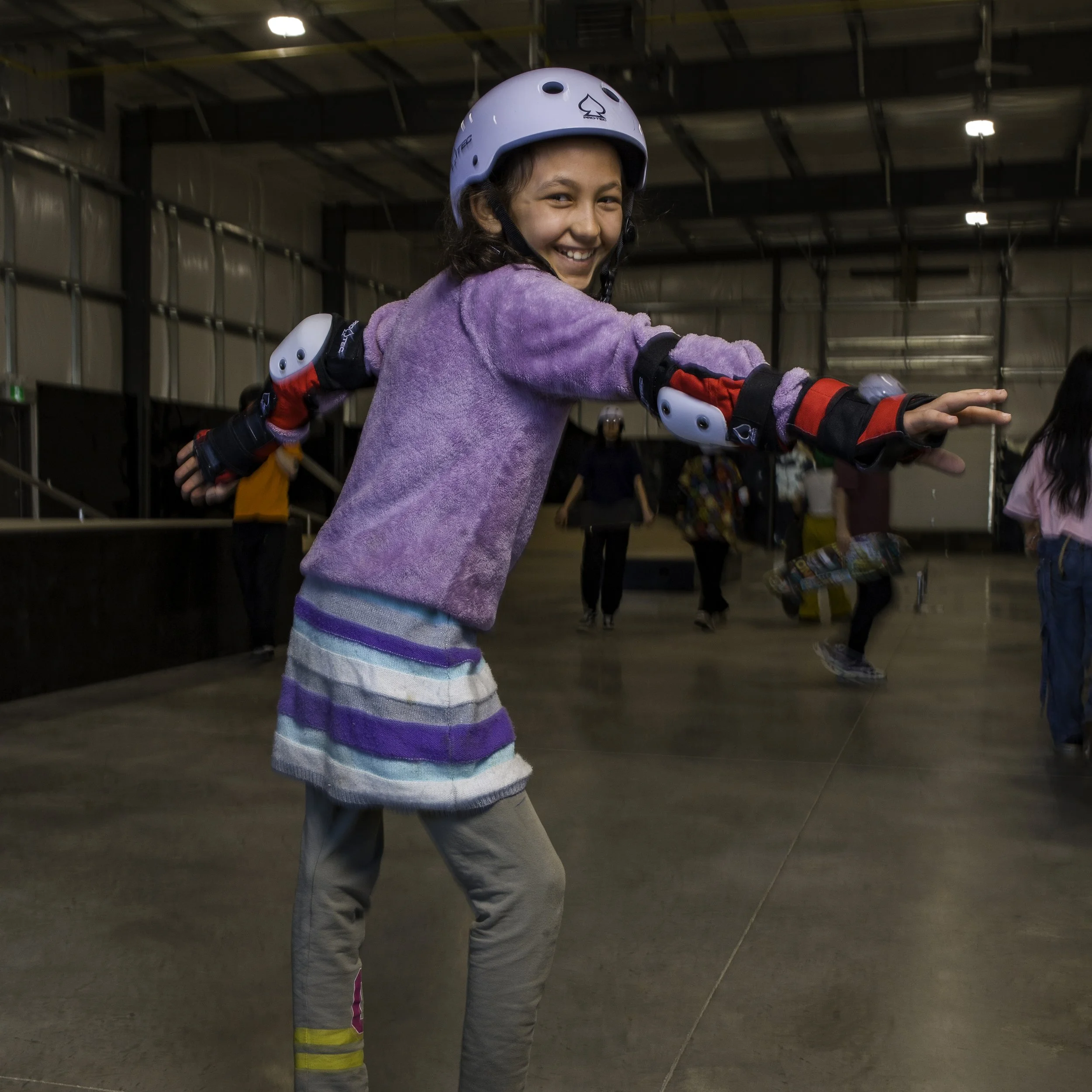 A young girl, one of Right To Skate's Afghan Team members, smiling and wearing a helmet, protective gear, and casual clothes, practicing roller skating indoors with other people in the background.