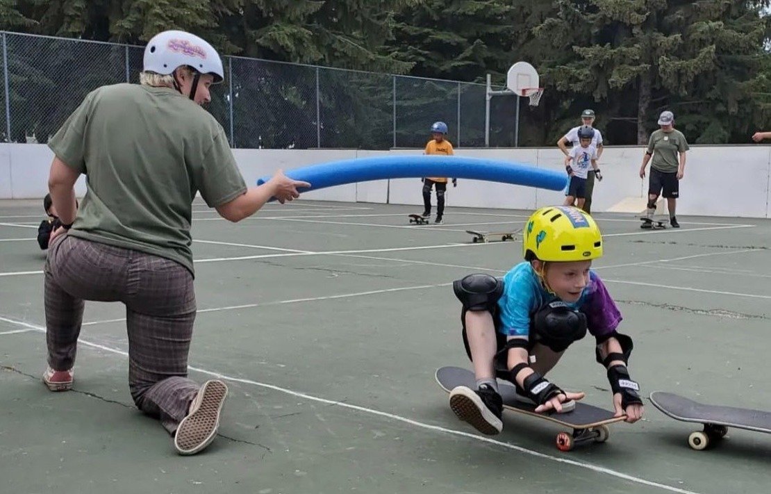 Girl in protective gear riding a skateboard on a skate rink while a woman holds a foam pool noodle overhead to help her. Several other skaters and a woman are in the background, all on skateboards at an outdoor rink surrounded by trees.