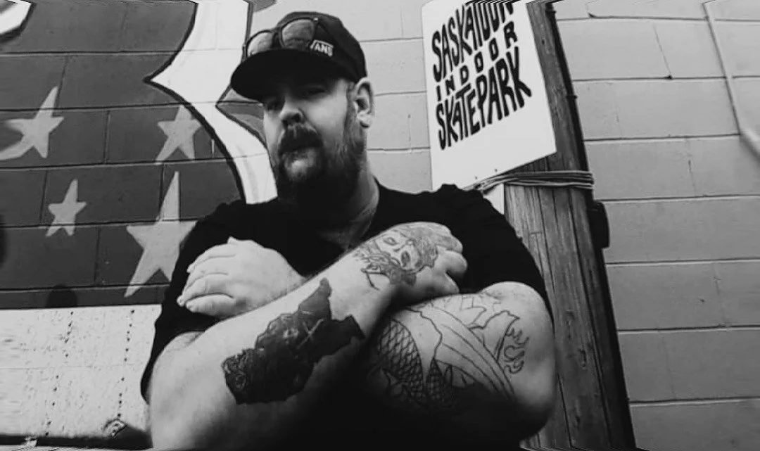 Right To Skate's Founder Joel Fortier, a man with a beard, wearing a baseball cap and sunglasses on top, crosses his arms with tattoos visible on his forearms, standing in front of a wall with a star mural.