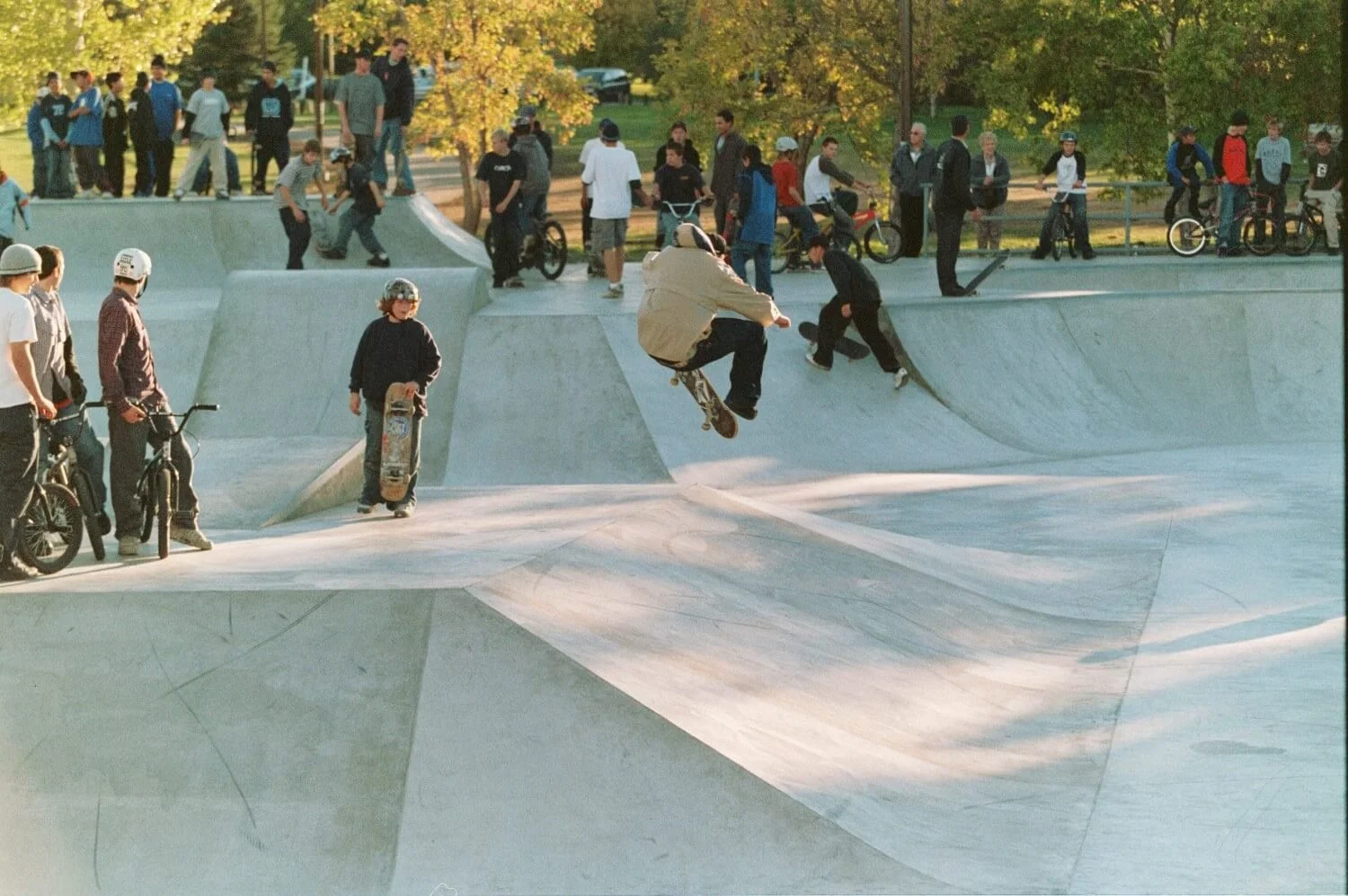 People skateboarding and riding bikes at a concrete skate park during the daytime, with trees and a grassy area in the background.