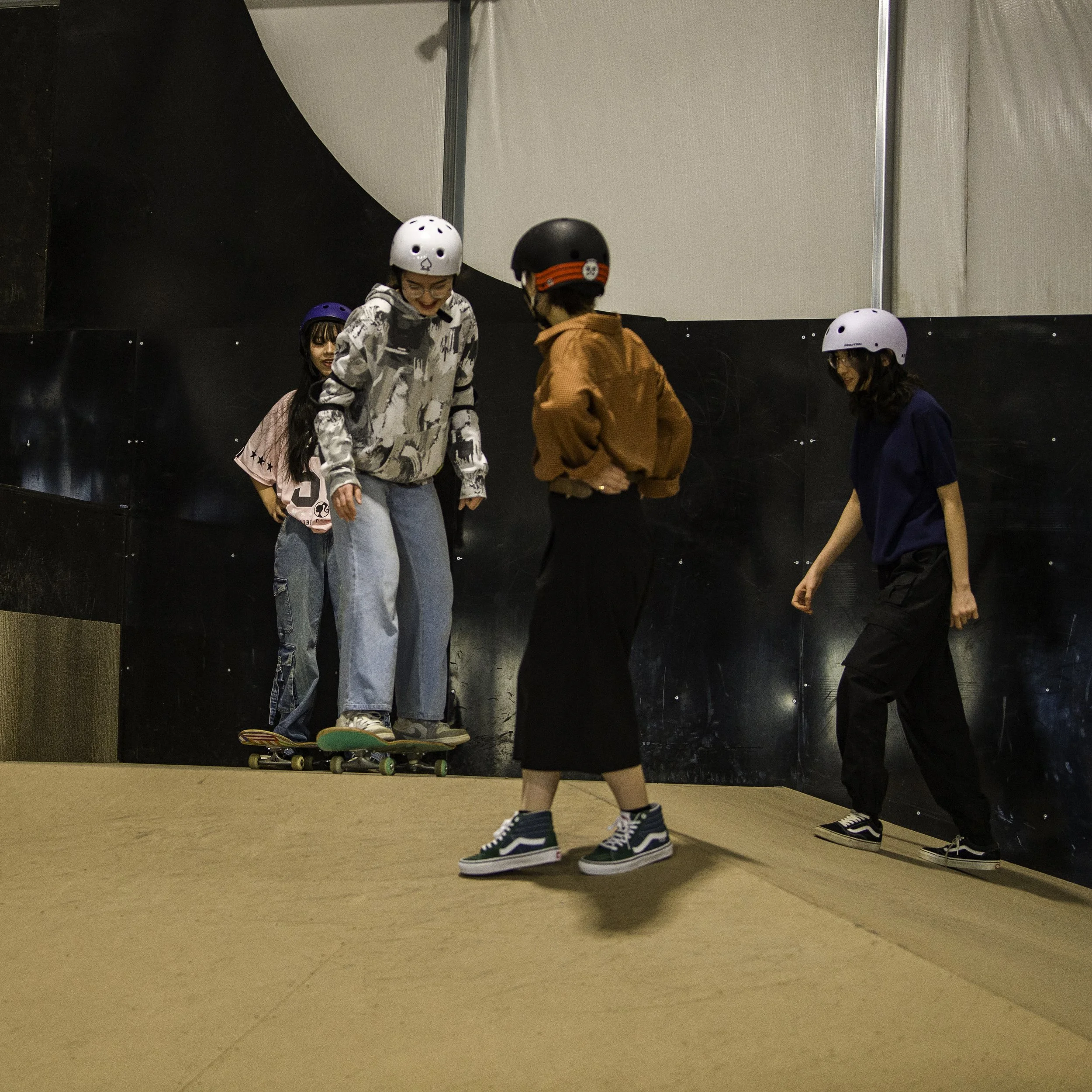 Four young people of Right To Skat's Afghan Skate Team wearing helmets at an indoor skatepark. One girl is riding a skateboard, while the others watch and talk.