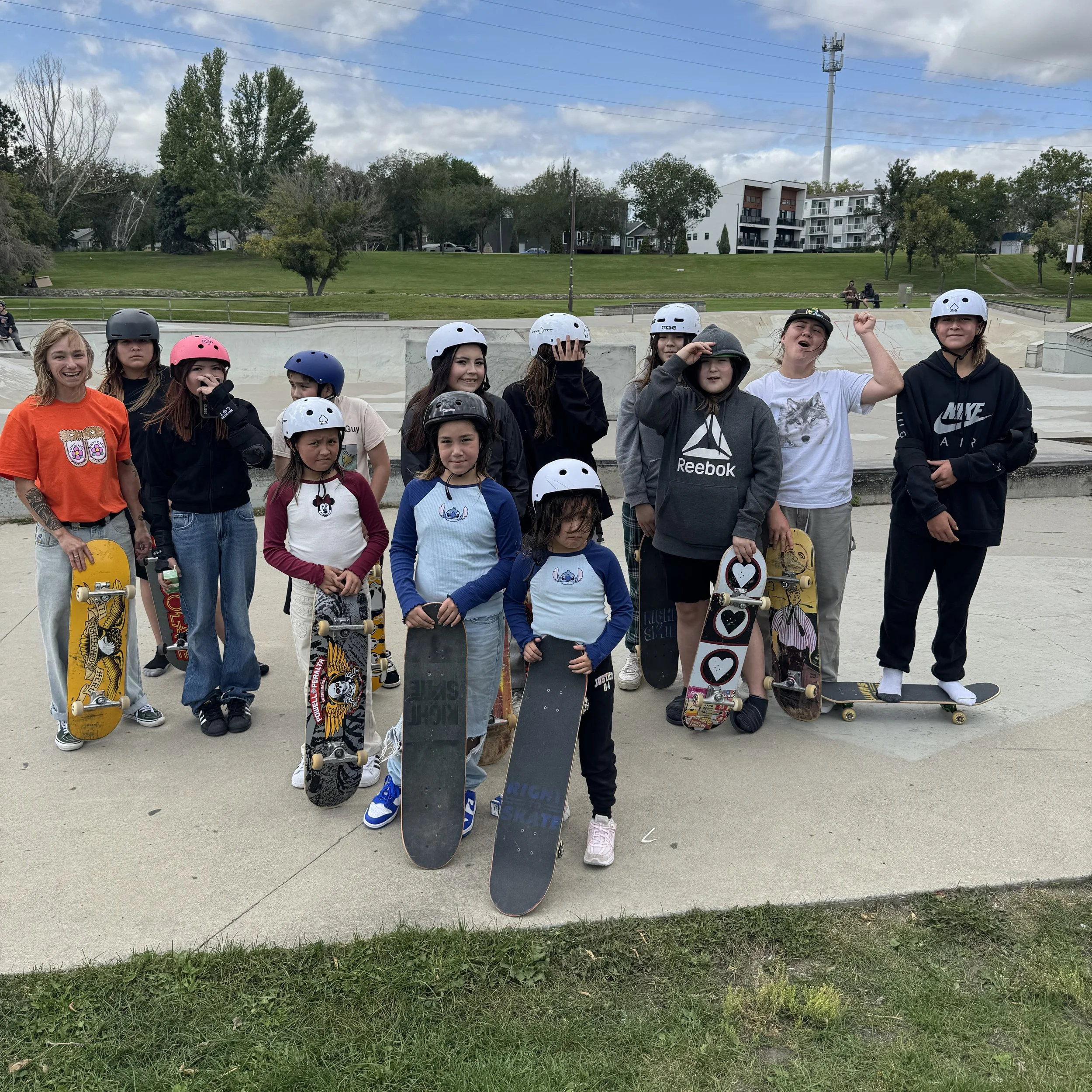 Three young girls at a skate park, two are holding skateboards and wearing helmets, and one is sitting on a concrete block holding Right To Skate certificates. The girls are smiling, and the scene is outdoors on a sunny day. 