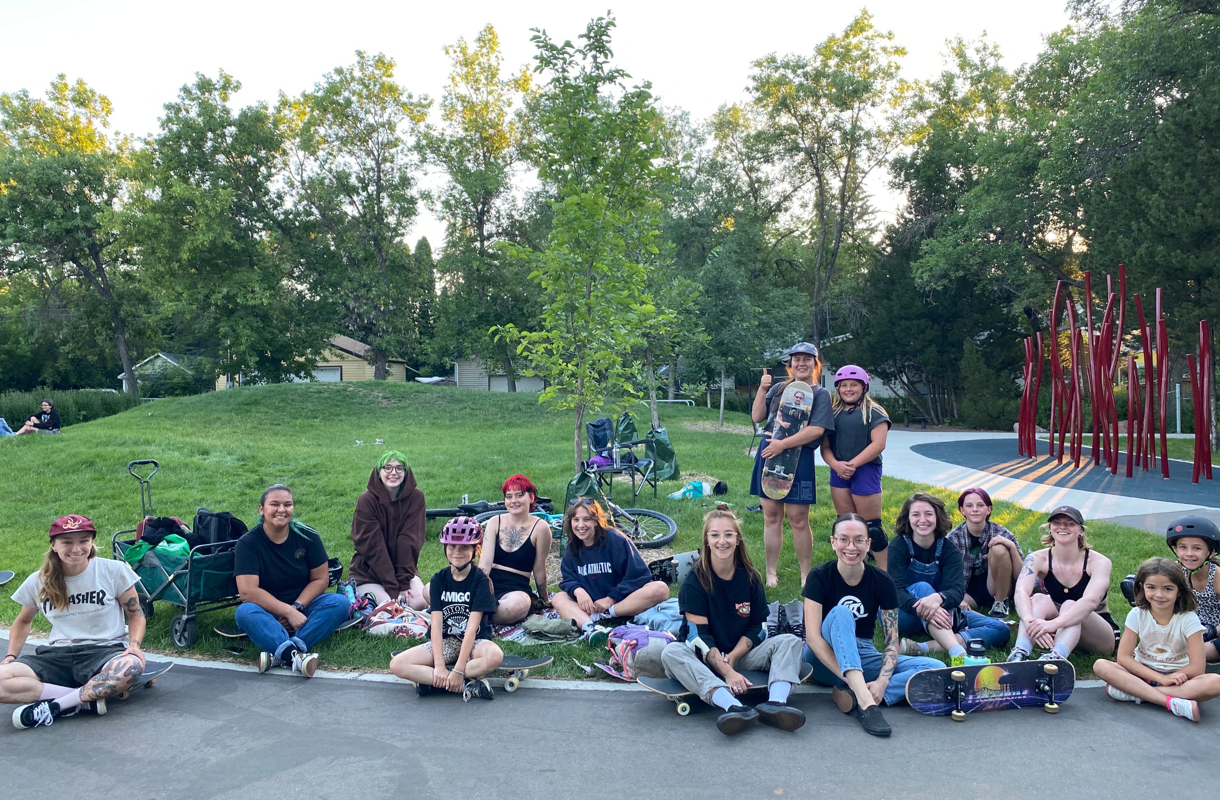 Group of people at a Right To Skate event, mostly women and girls, sitting and standing outdoors on a grassy area with skateboards, bikes, and backpacks, smiling, with trees and a playground structure in the background.