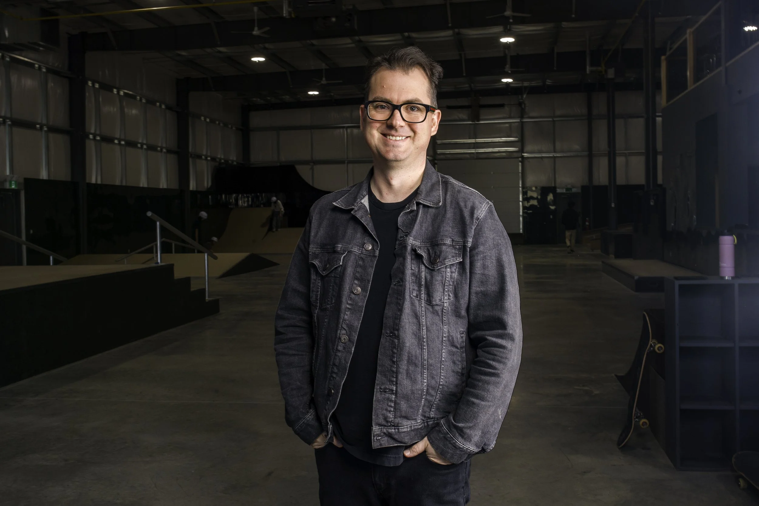 Right To Skate's Director Dallas Tetarenko, a young man in a black denim jacket and glasses smiling inside an indoor skate park with ramps and skateboarders in the background.