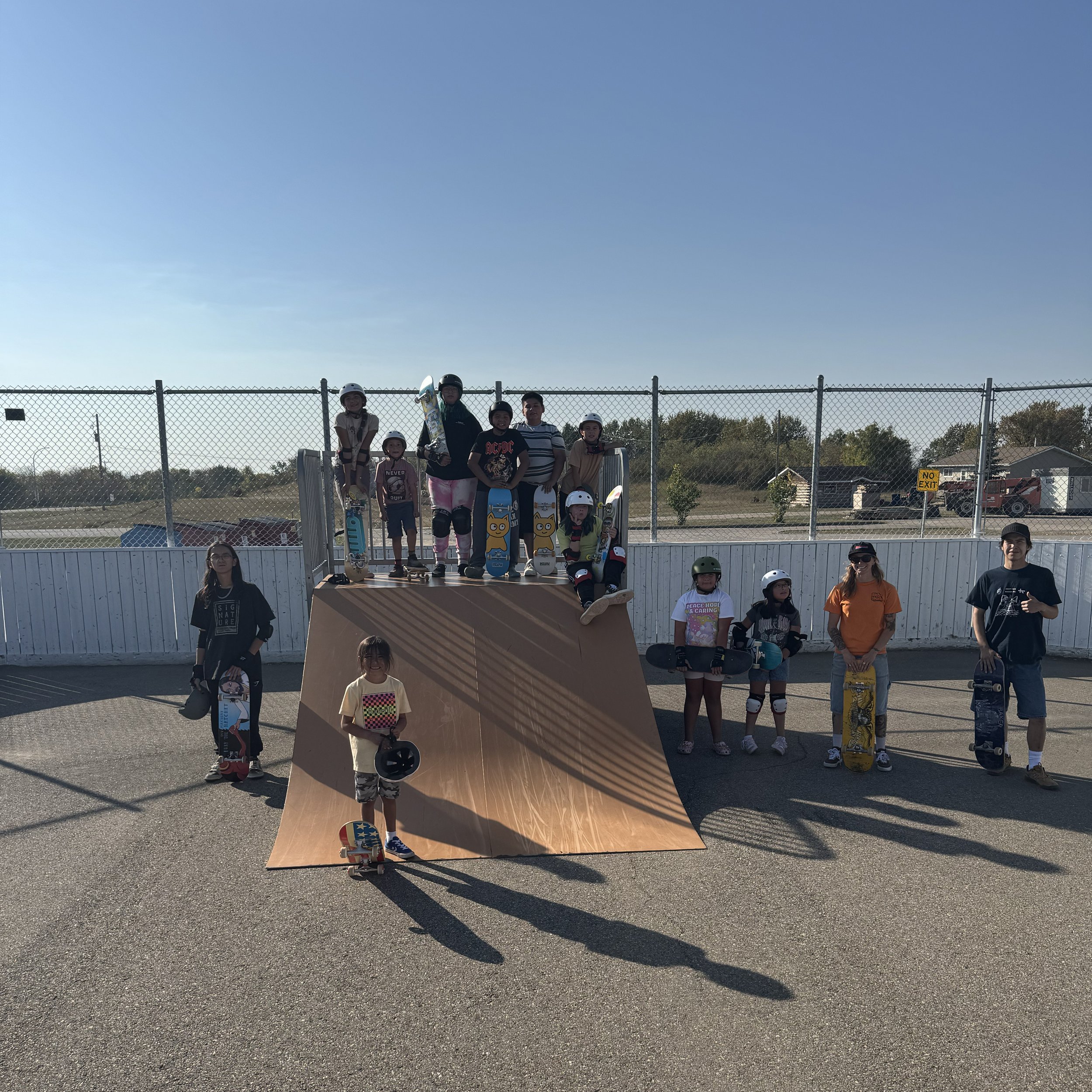 Indigenous children and teenagers in a Right To Skate class at a skate park, wearing helmets and holding skateboards, with some standing on a ramp and others on the ground, under a clear blue sky.