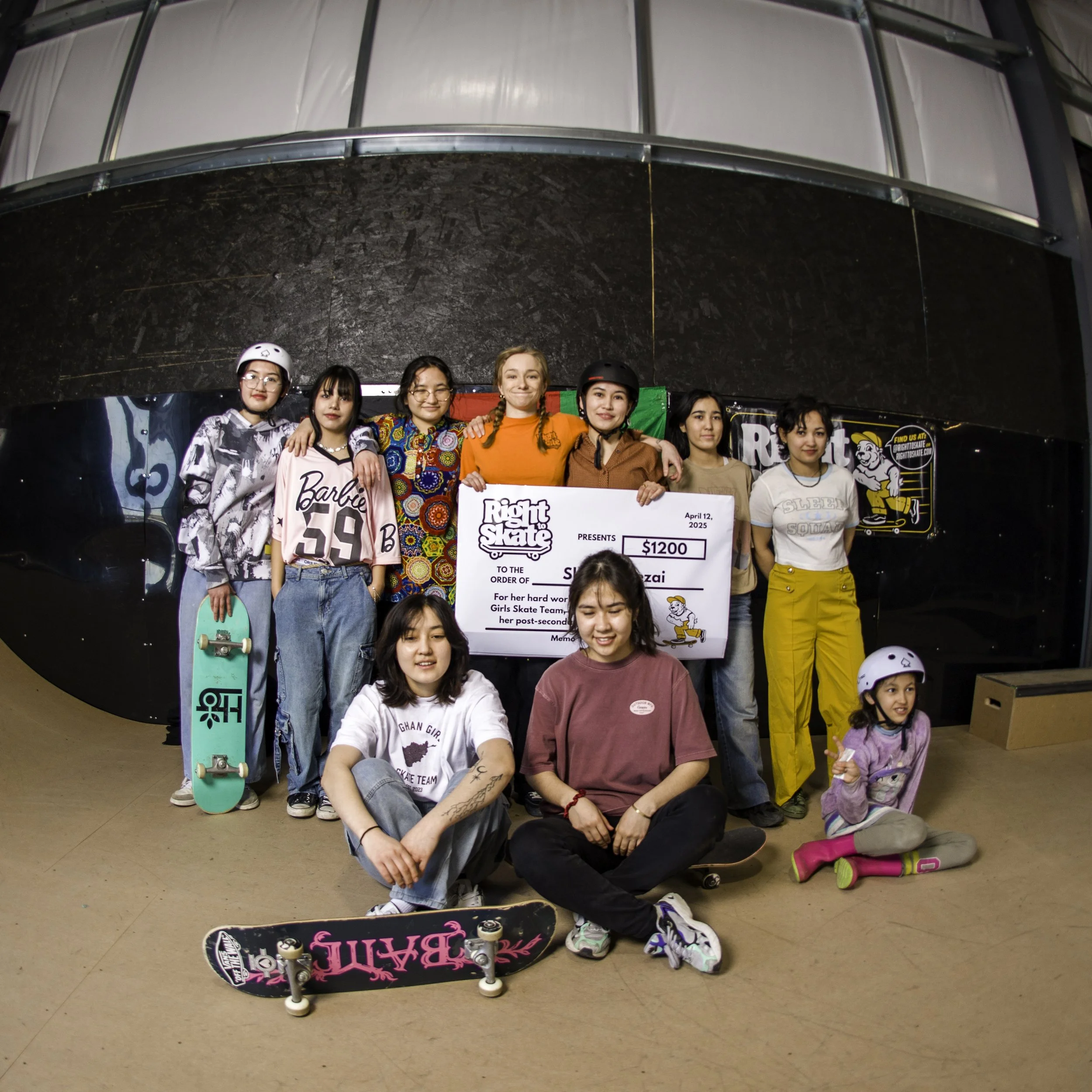 Right To Skate's Afghan Skate Team celebrating at a skateboarding event, holding a giant check for $1200, with skateboards and wearing helmets, in an indoor skate rink.