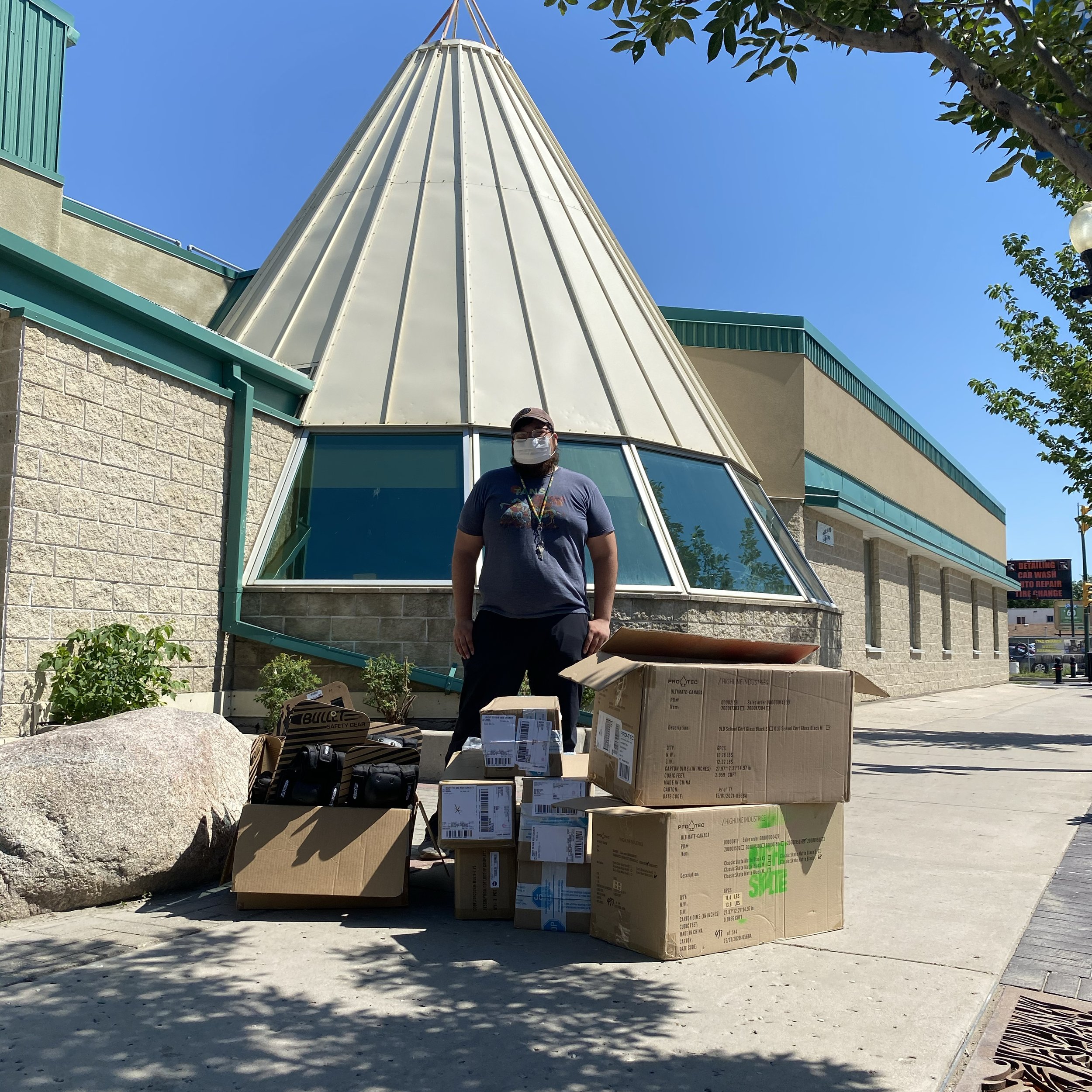 Person standing outdoors wearing a mask, surrounded by Right To Skate boxes and bags, in front of a building with a teepee-shaped roof and large windows.