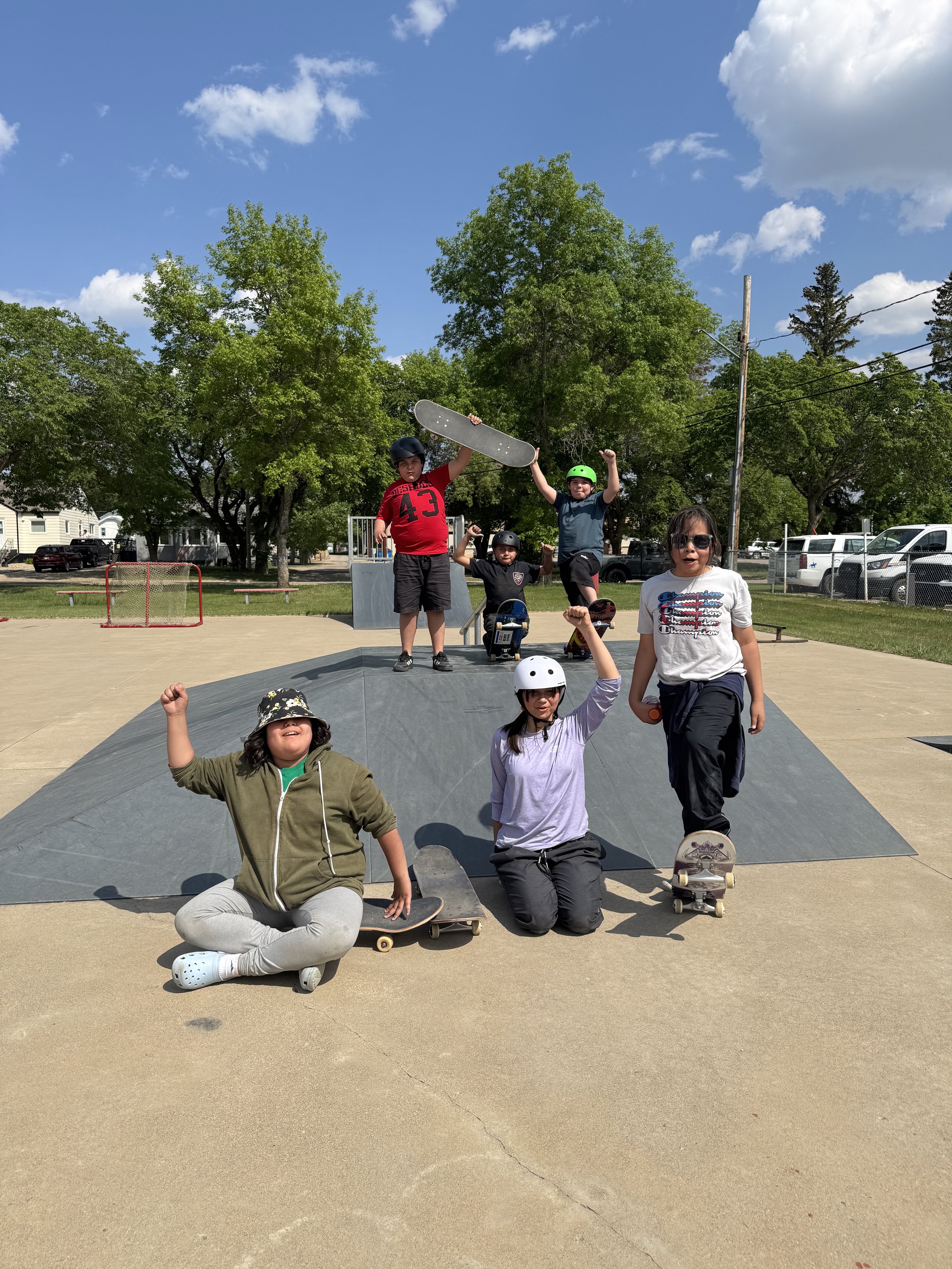 Group of Indigenous children posing on and around a skate park ramp outdoors on a sunny day. Some children are holding Right To Skate skateboards, wearing helmets, and making celebratory gestures.