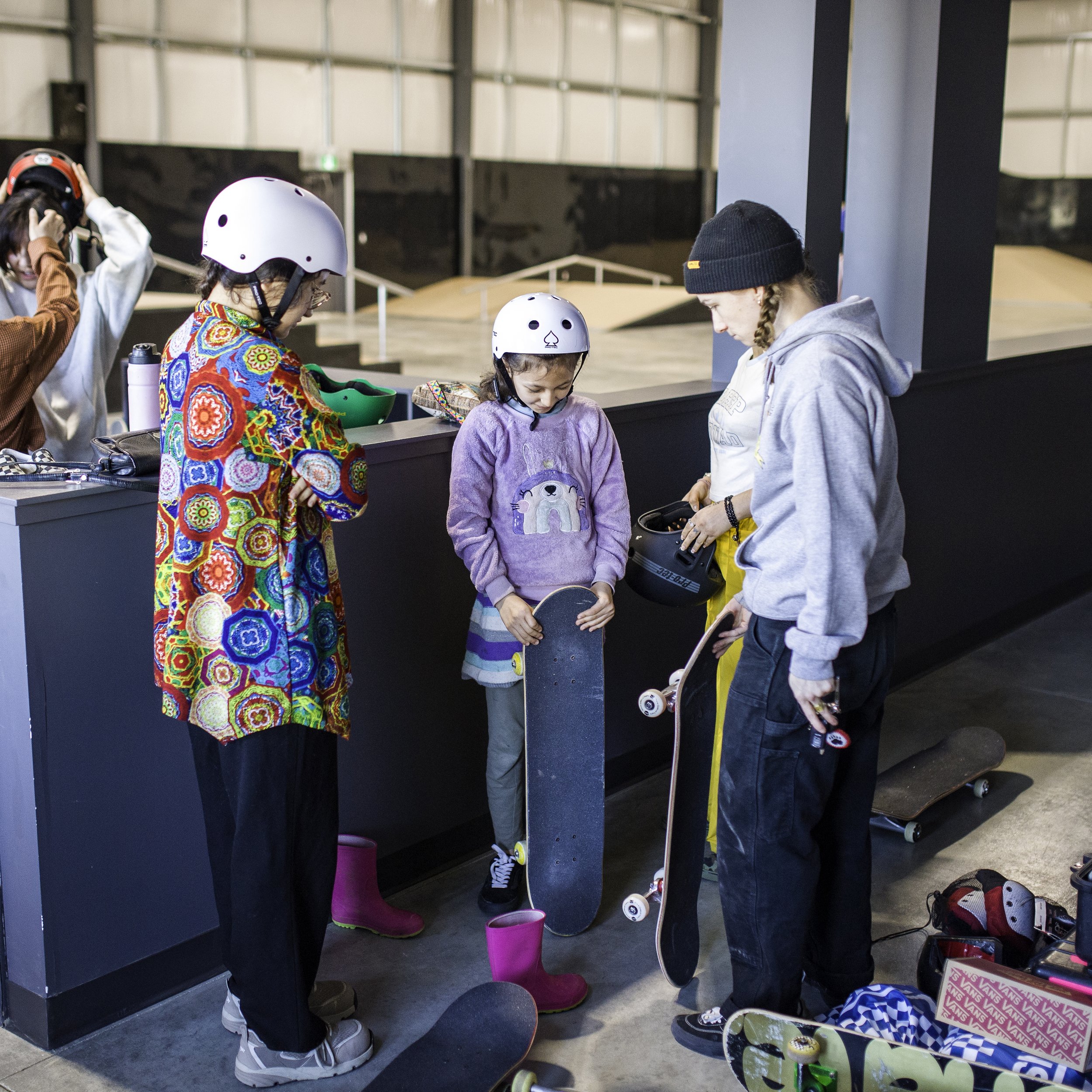 A group of young girls in Right To Skate's Afghan Skate Team and a woman wearing helmets, holding skateboards, and talking inside a skatepark. The activity is about skateboarding lessons or practice.