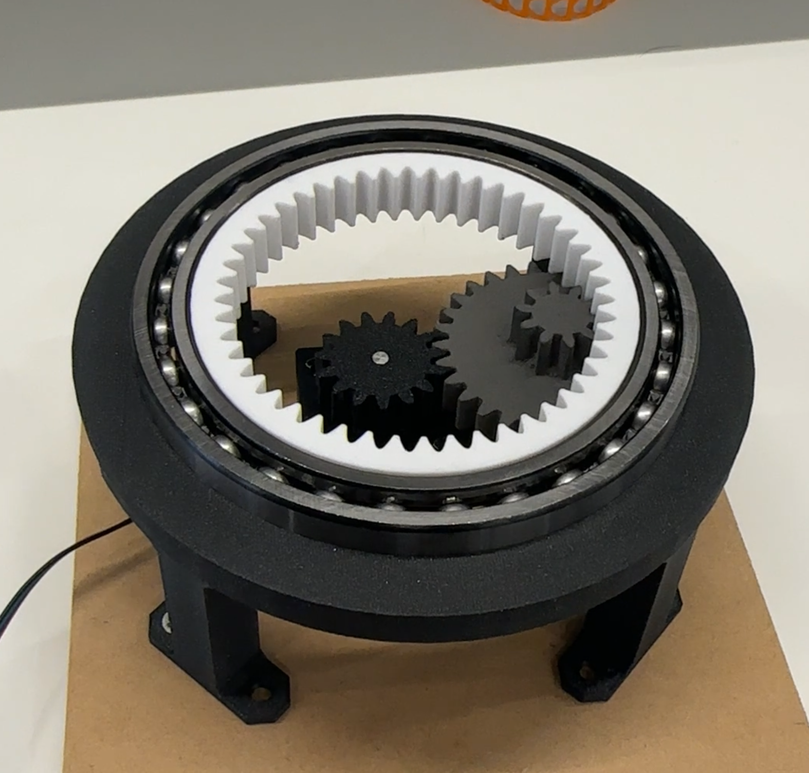 A planetary gear system with multiple black gears inside a large circular housing, mounted on a black stand.